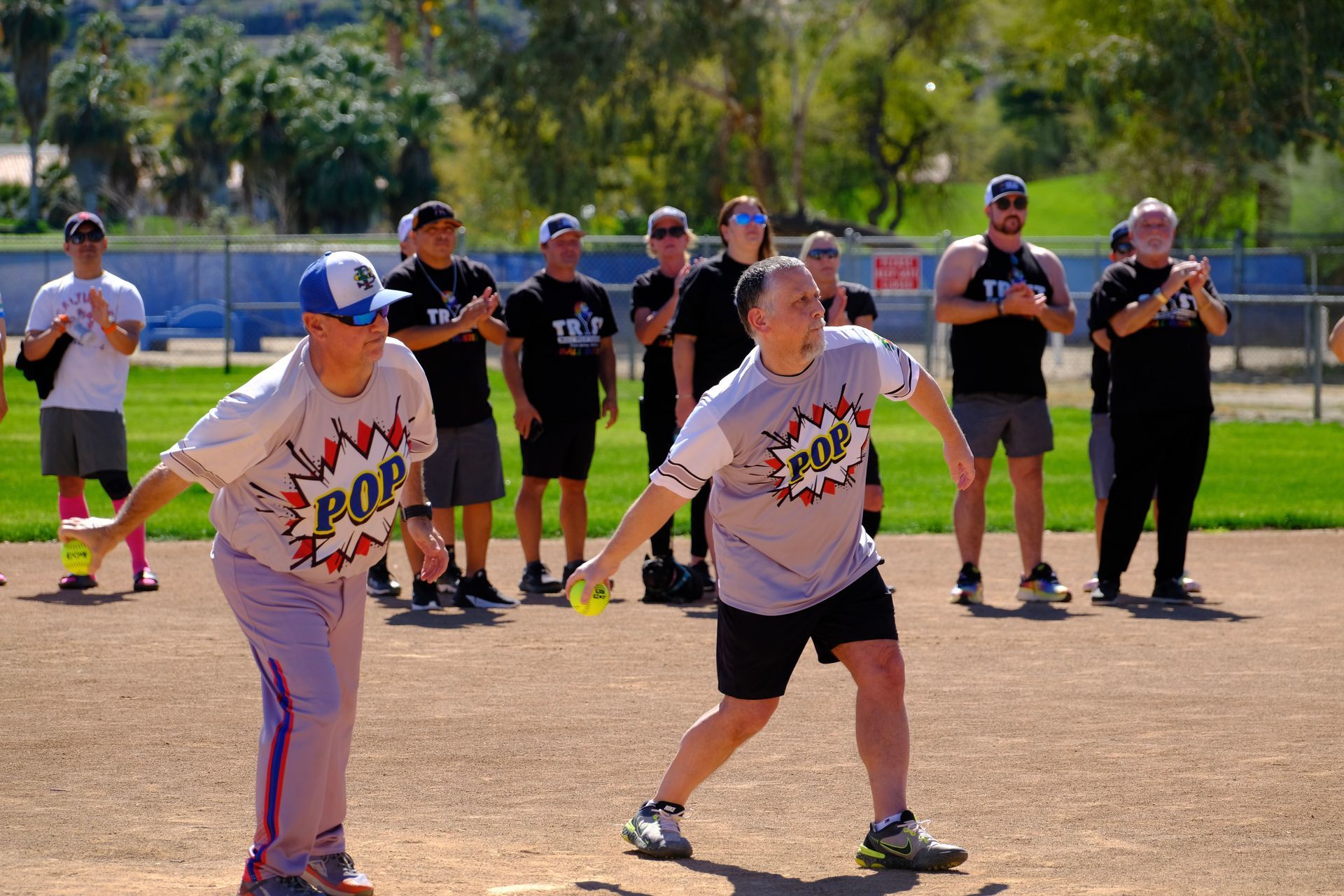 Two softball players in gray shirts at bat, with teammates watching on a sunny field.