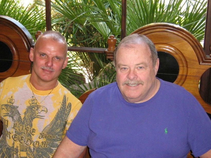 Two men sitting together outdoors, one in a yellow shirt and one in a purple shirt, with plants behind them