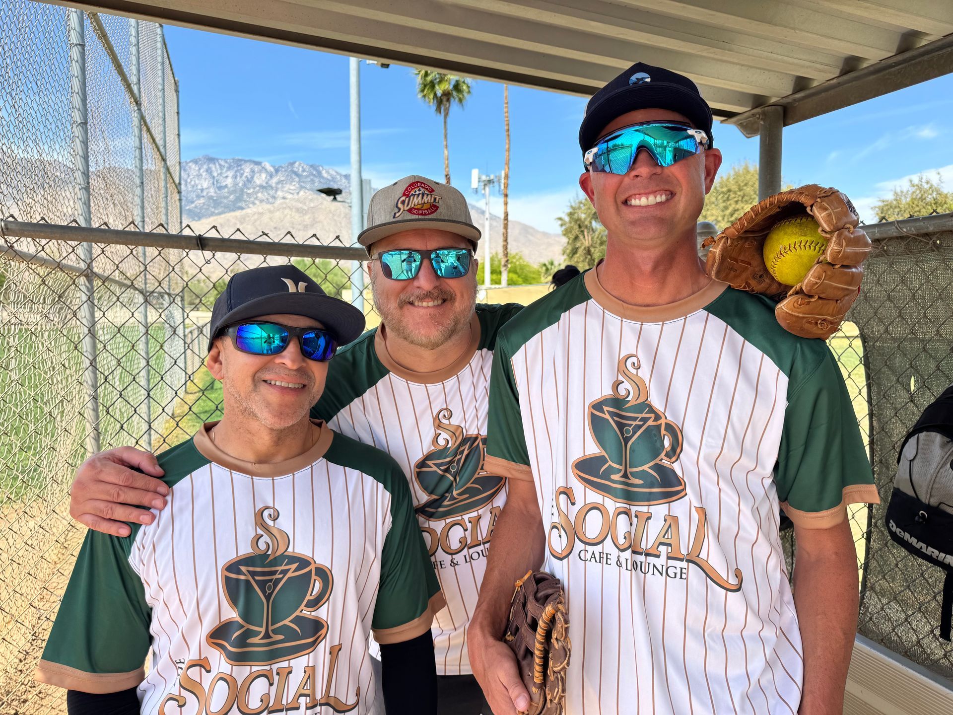 Three baseball players in white and green uniforms smiling under a dugout roof, one holding a glove with a ball.