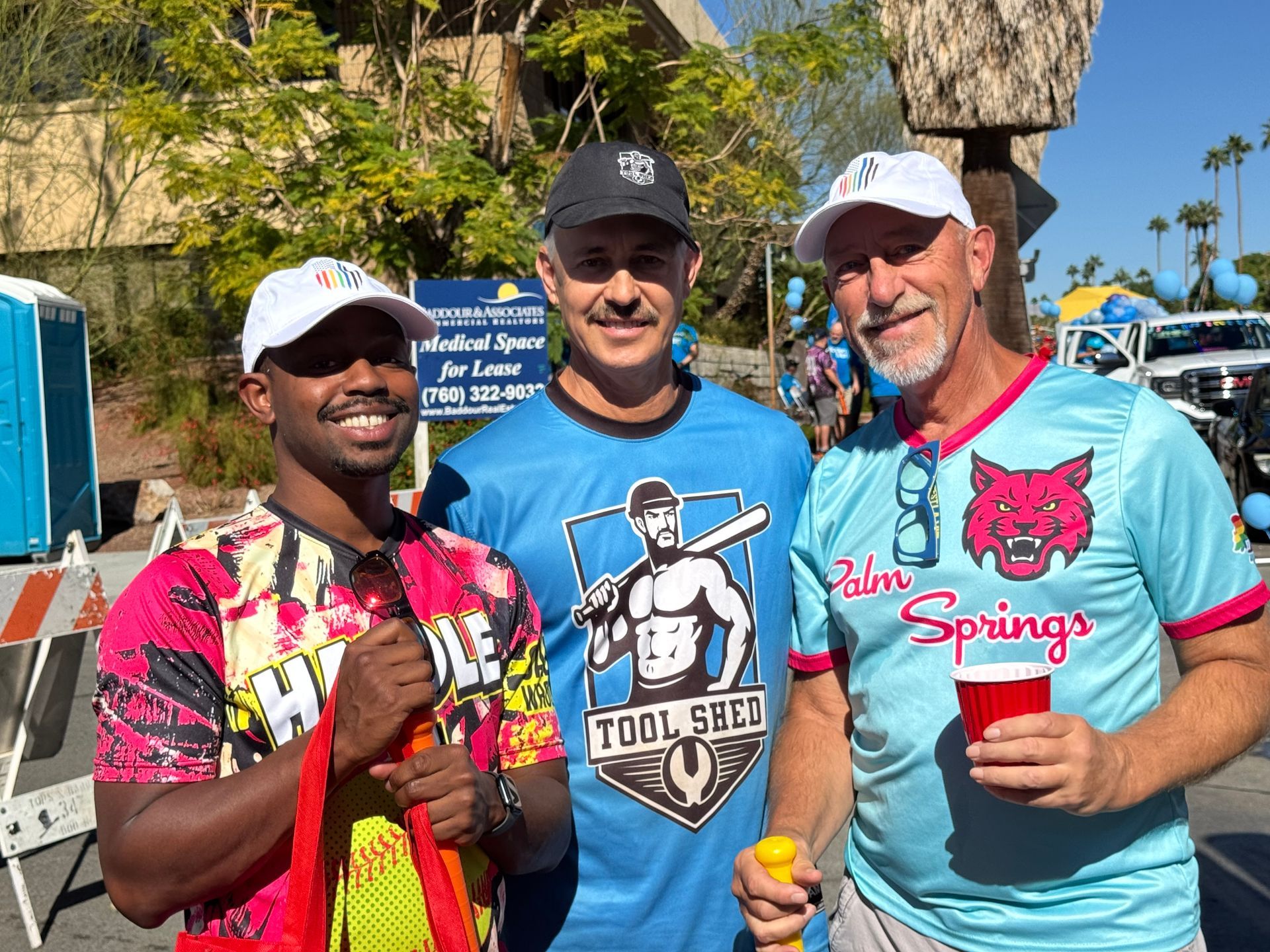 Three smiling people hold drinks outdoors at a colorful event, wearing casual sporty shirts and caps.