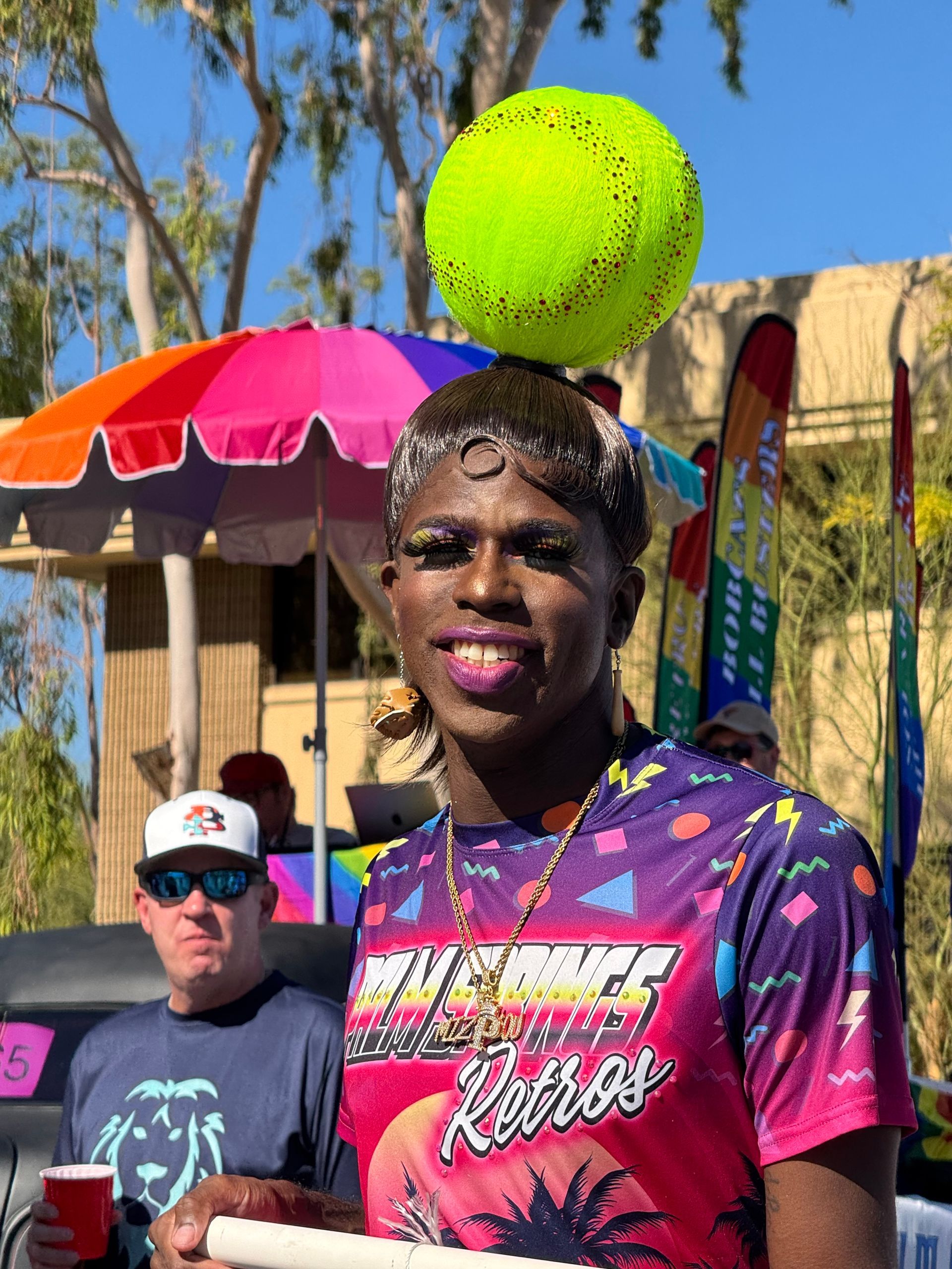 Person in colorful shirt at outdoor event with a large tennis ball prop overhead.