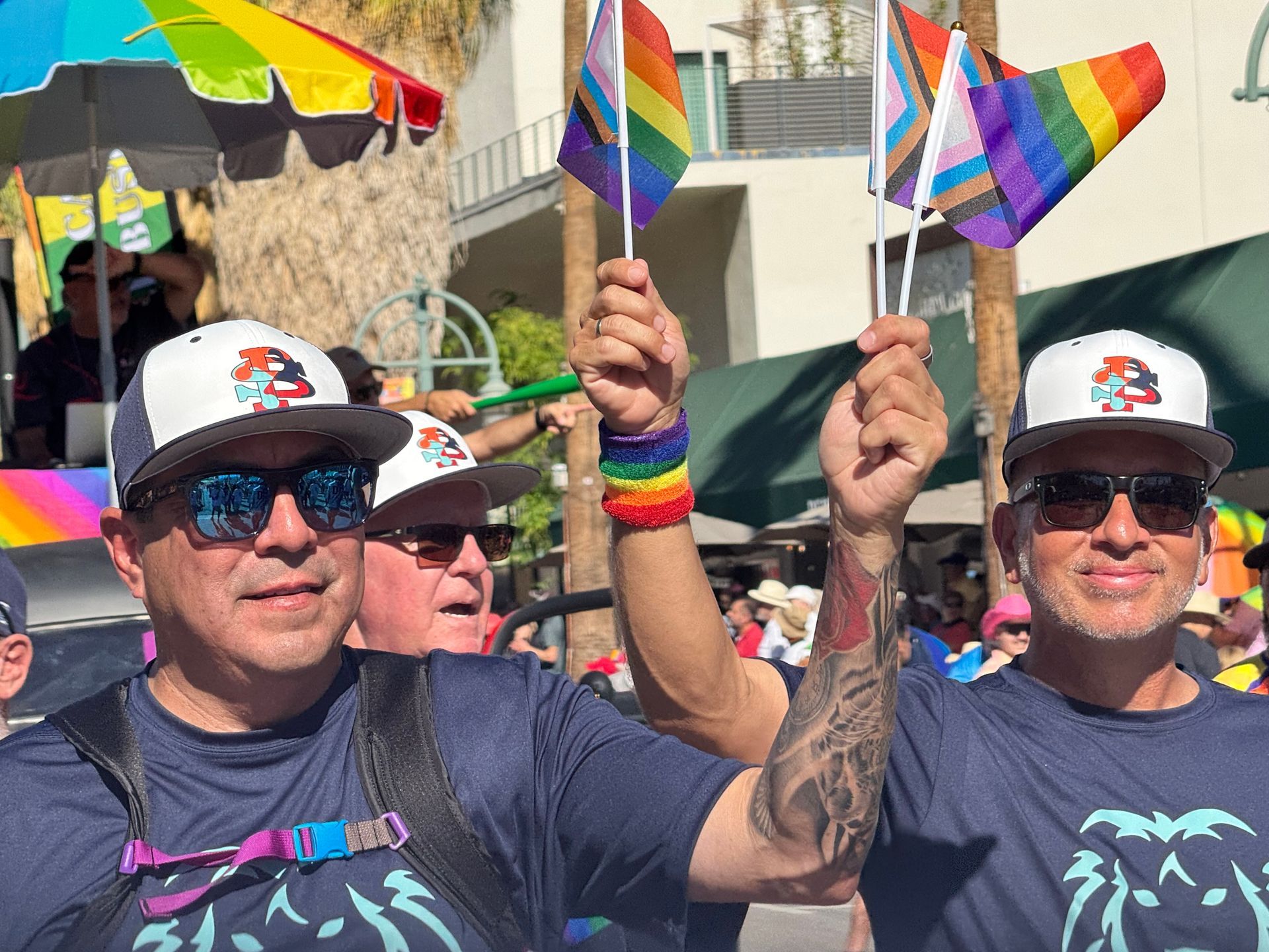 Crowd marching with rainbow flags at a Pride parade, two people in sunglasses in front.
