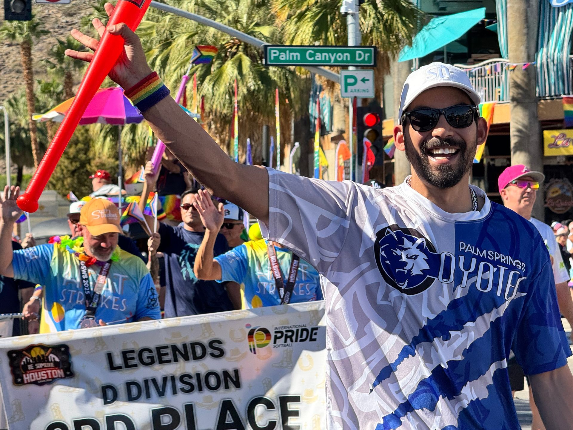 Pride parade crowd with flags and a smiling person in sunglasses waving a red baton on a palm-lined street
