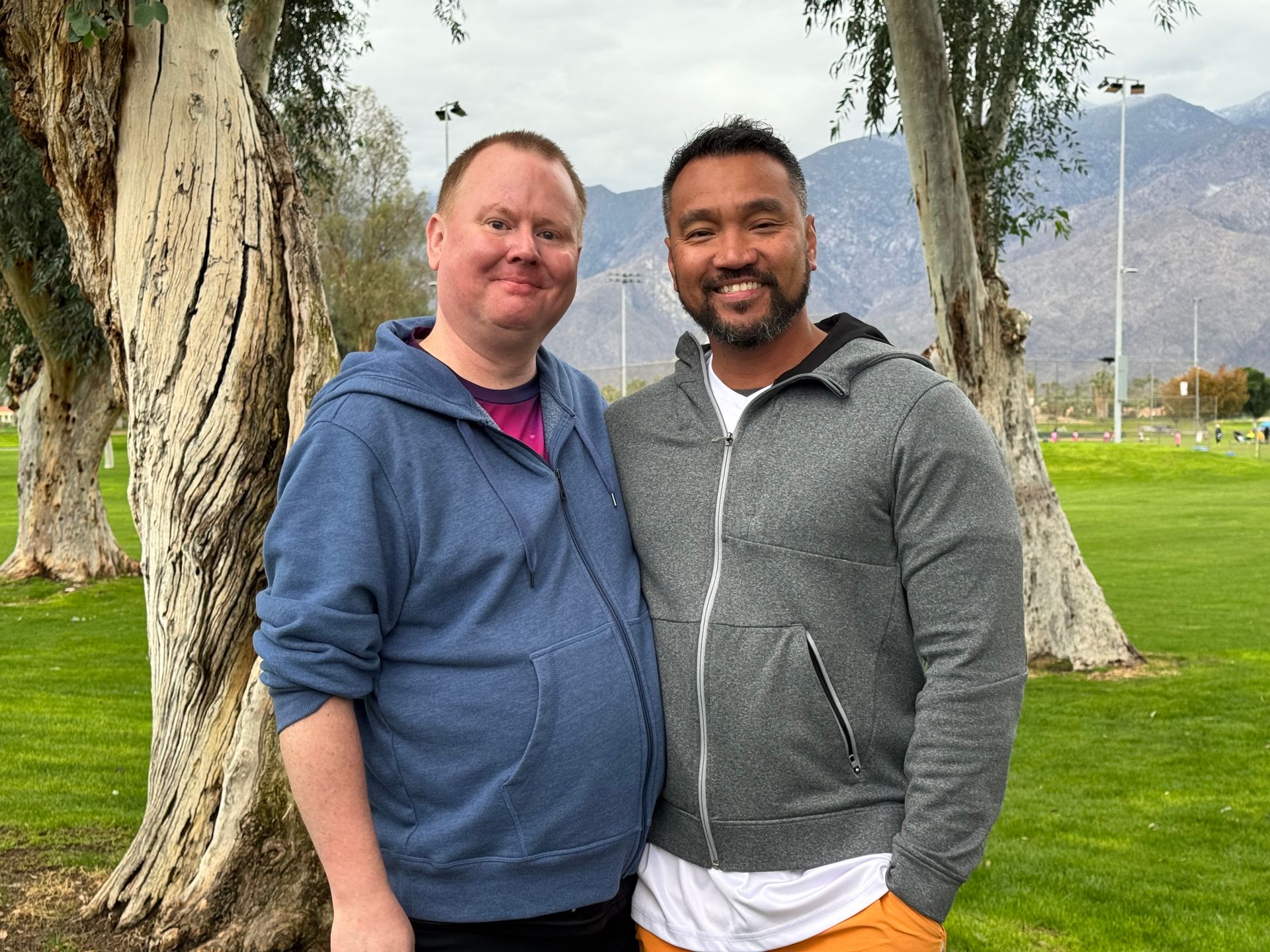 Two men standing together in a park, smiling, with trees and mountains in the background.