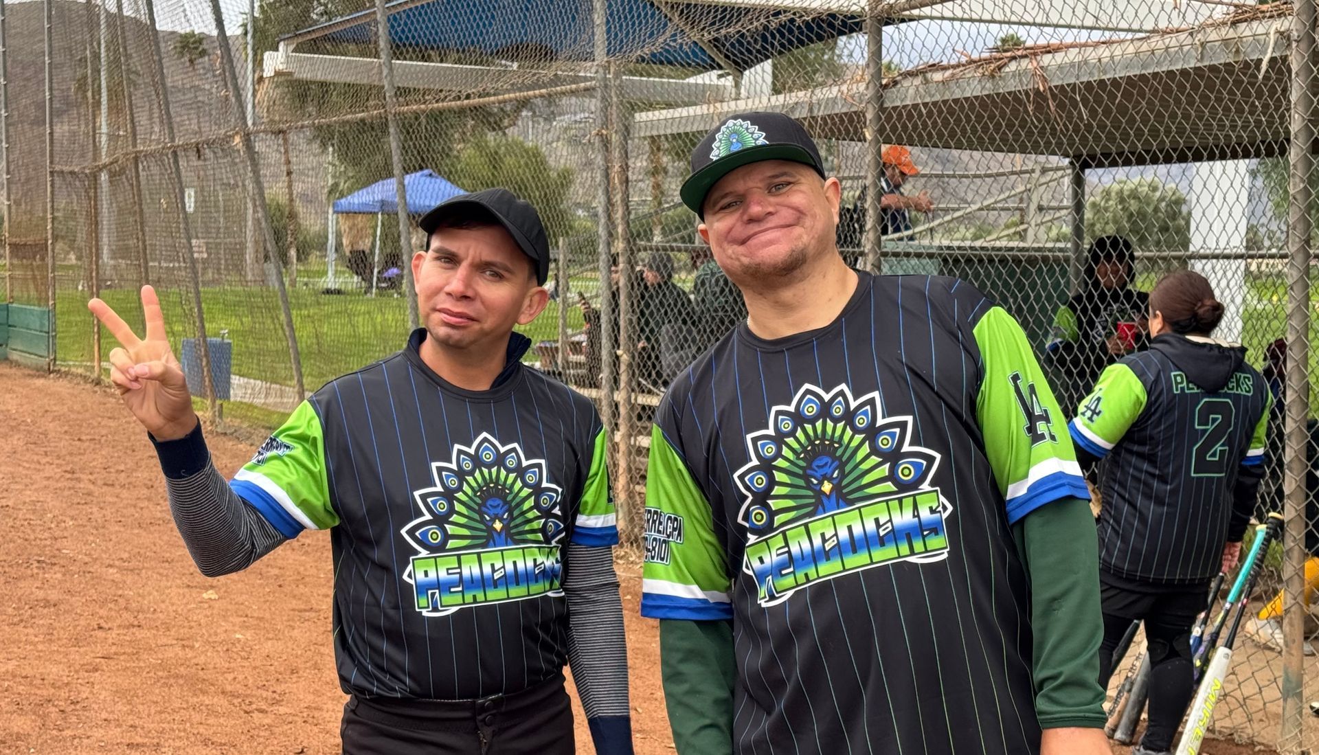 Two men in matching black-and-green sports jerseys pose at a baseball field, one flashing a peace sign.