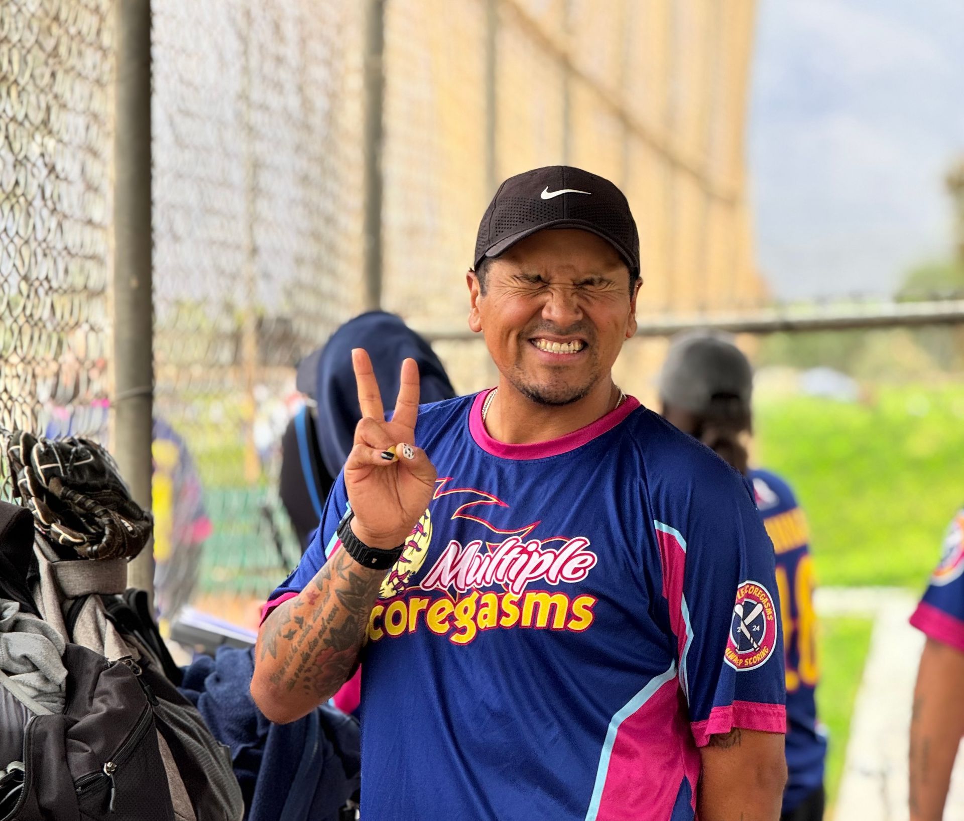 Smiling man in a black cap and blue jersey flashes a peace sign at a baseball field fence.