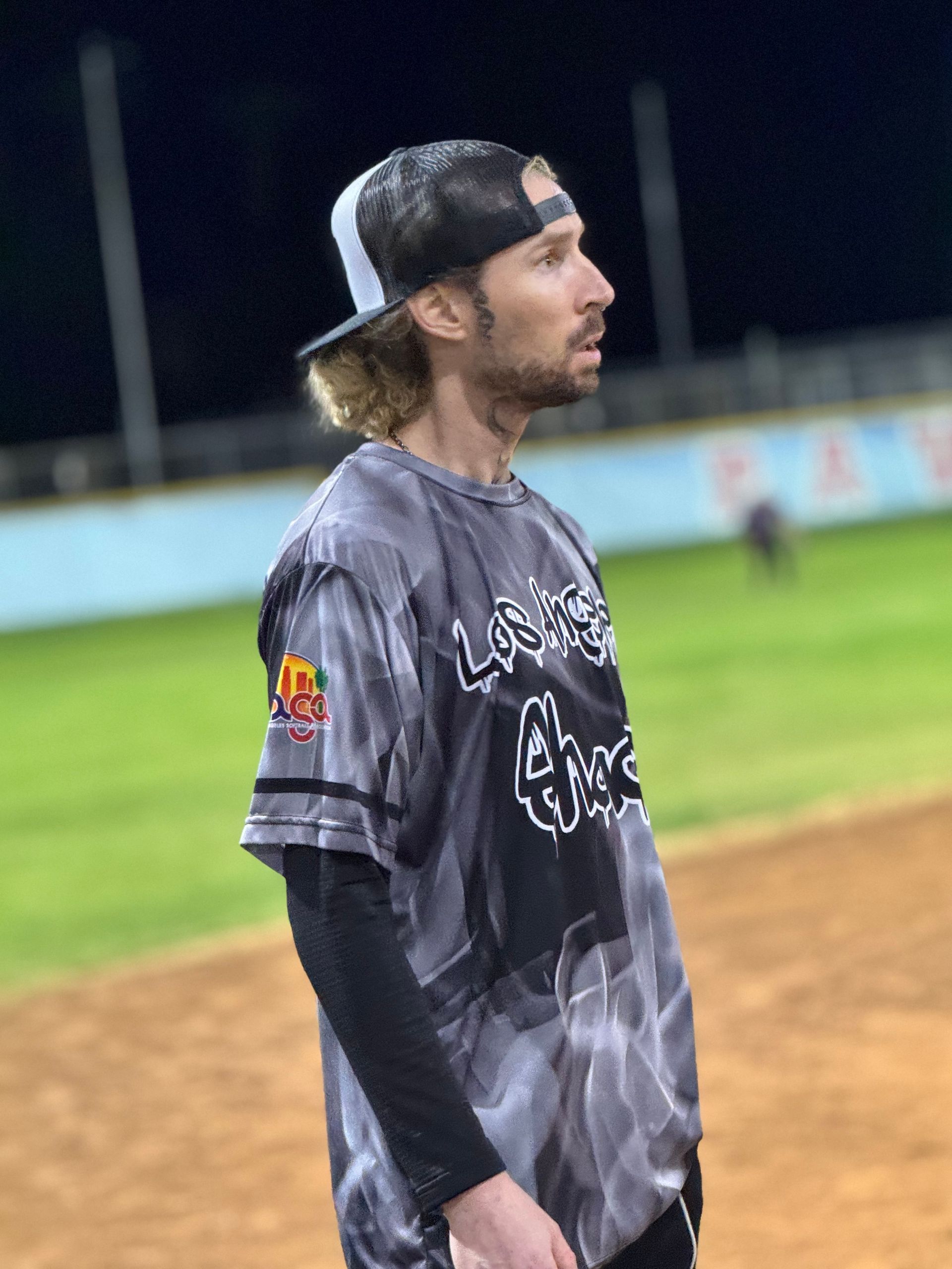 Baseball player in a gray jersey and black cap stands on the field at night, looking to the side.