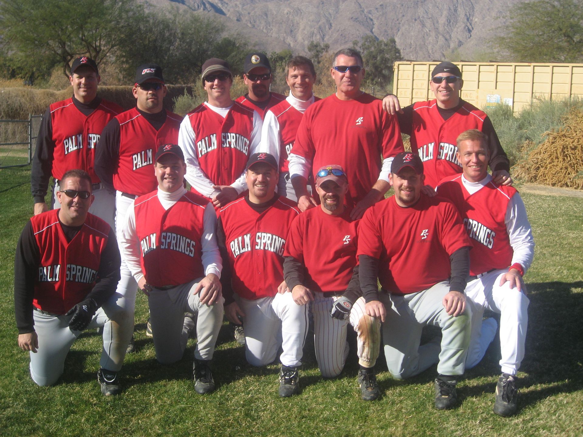 Group of baseball players in red jerseys posing outdoors on grass, with mountains in the background