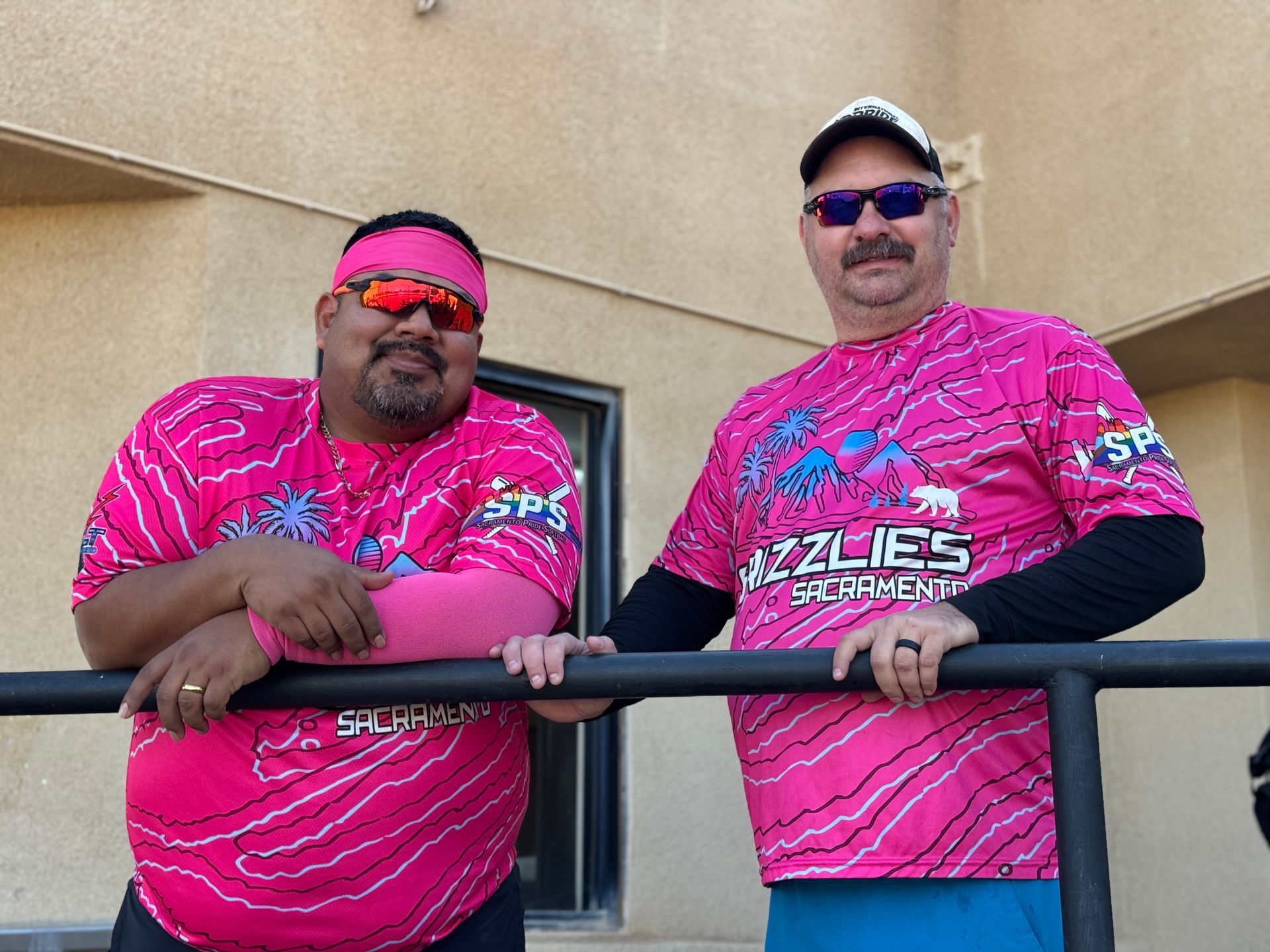 Two men in pink jerseys and sunglasses lean on a railing outdoors.