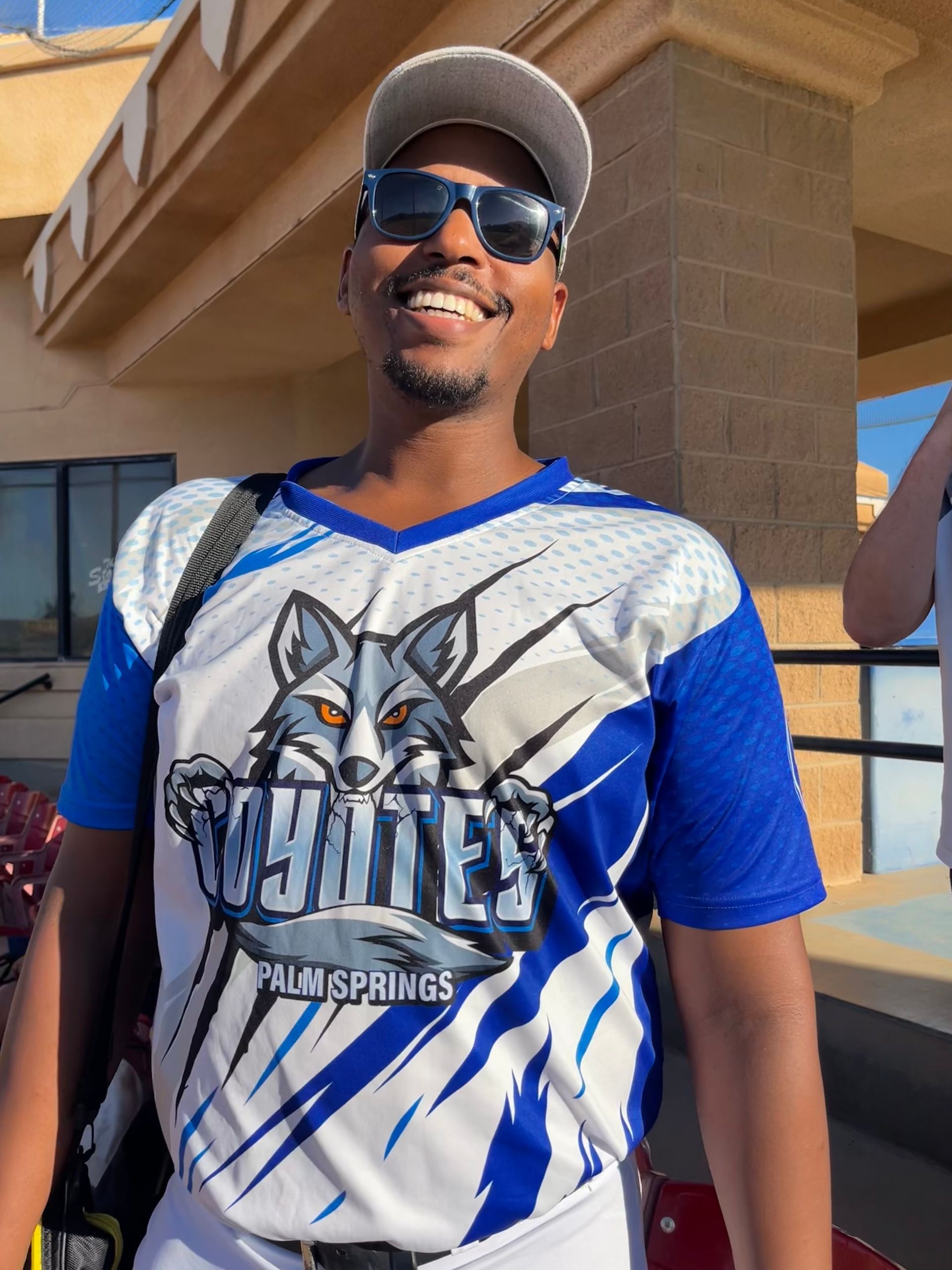 Man wearing sunglasses and a blue sports jersey, smiling outdoors beside a building.