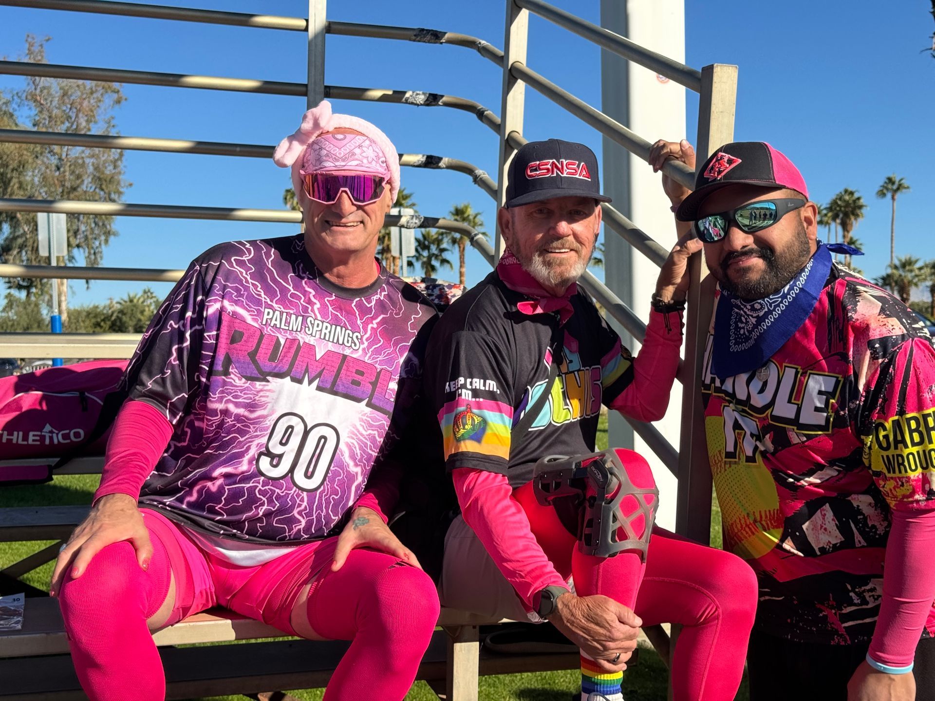 Three people in bright pink cycling gear sitting on a bench outdoors, smiling at the camera.