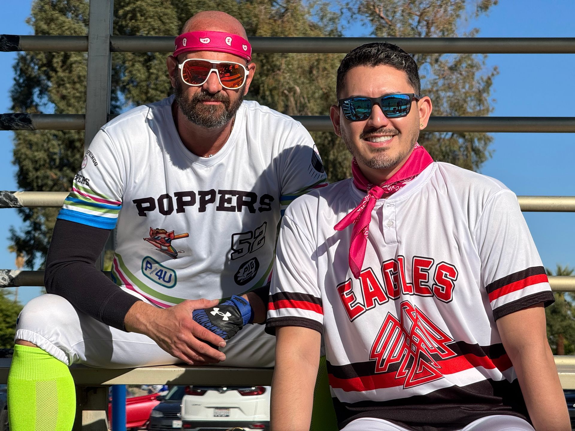 Two men in baseball uniforms and sunglasses pose outdoors on bleachers under a blue sky.