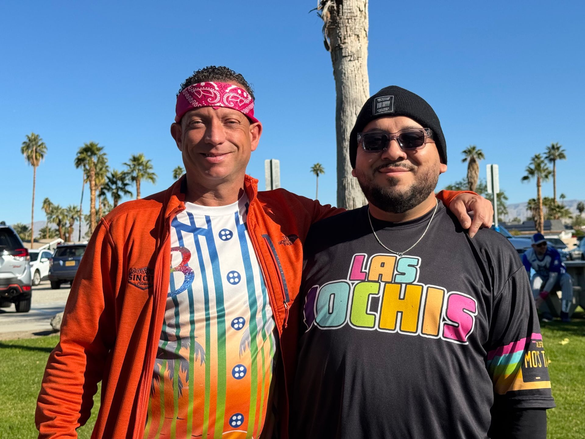 Two people standing outdoors in bright sun, wearing colorful shirts and smiling at the camera.