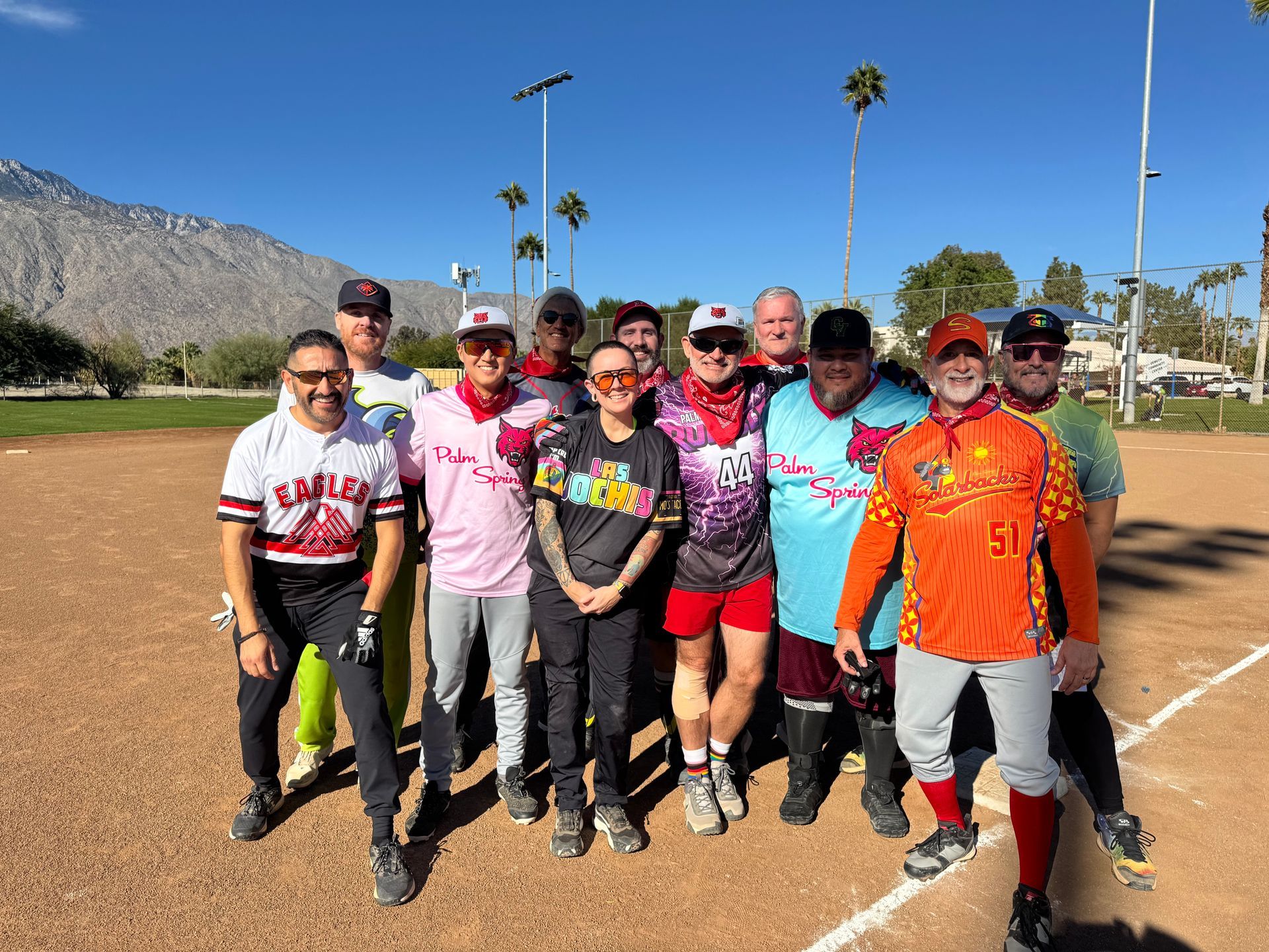 Group of cyclists posing on a baseball field, wearing colorful jerseys and helmets under a sunny sky