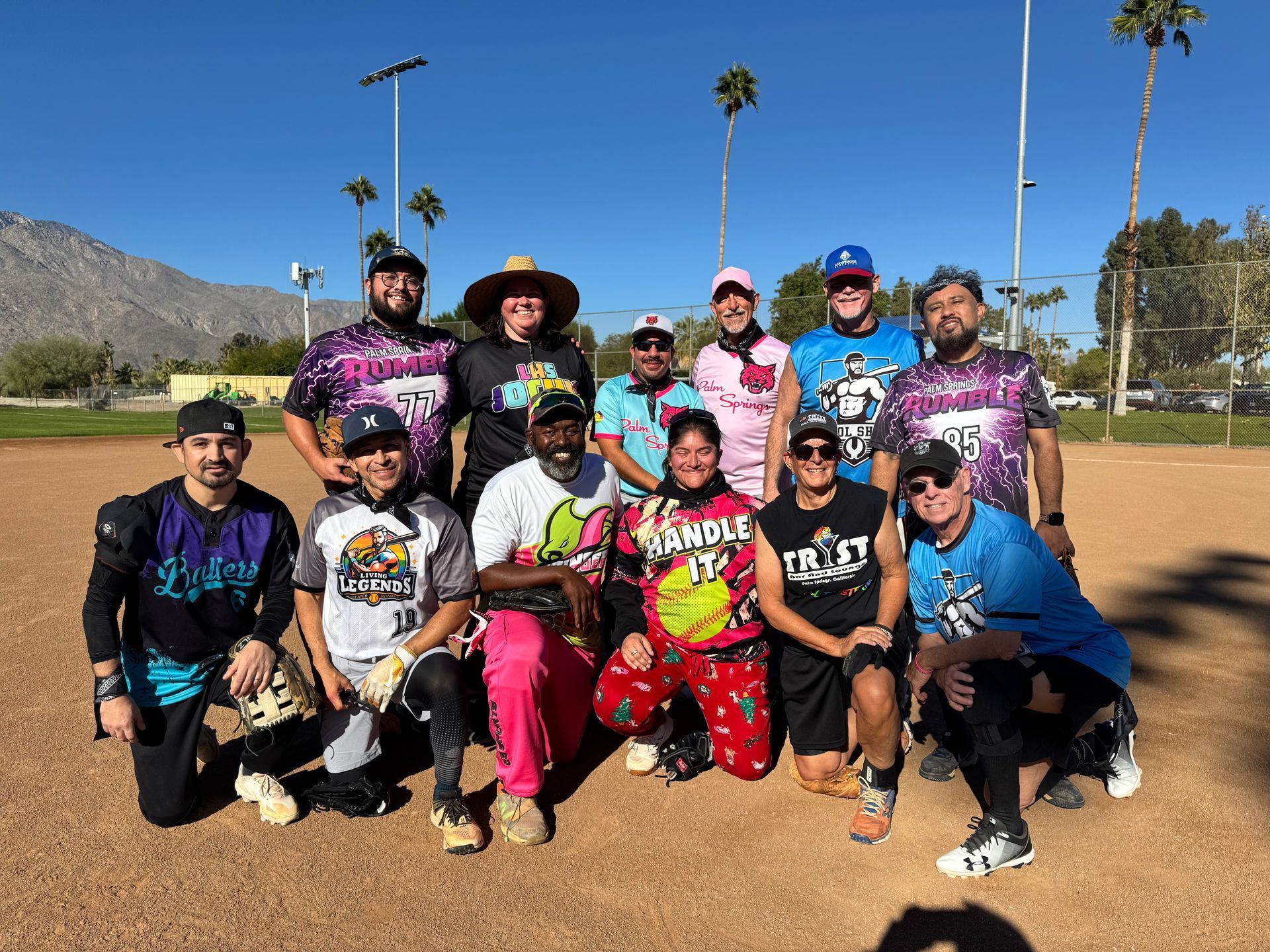 Group of people posing together on a softball field in bright athletic uniforms under a clear blue sky.