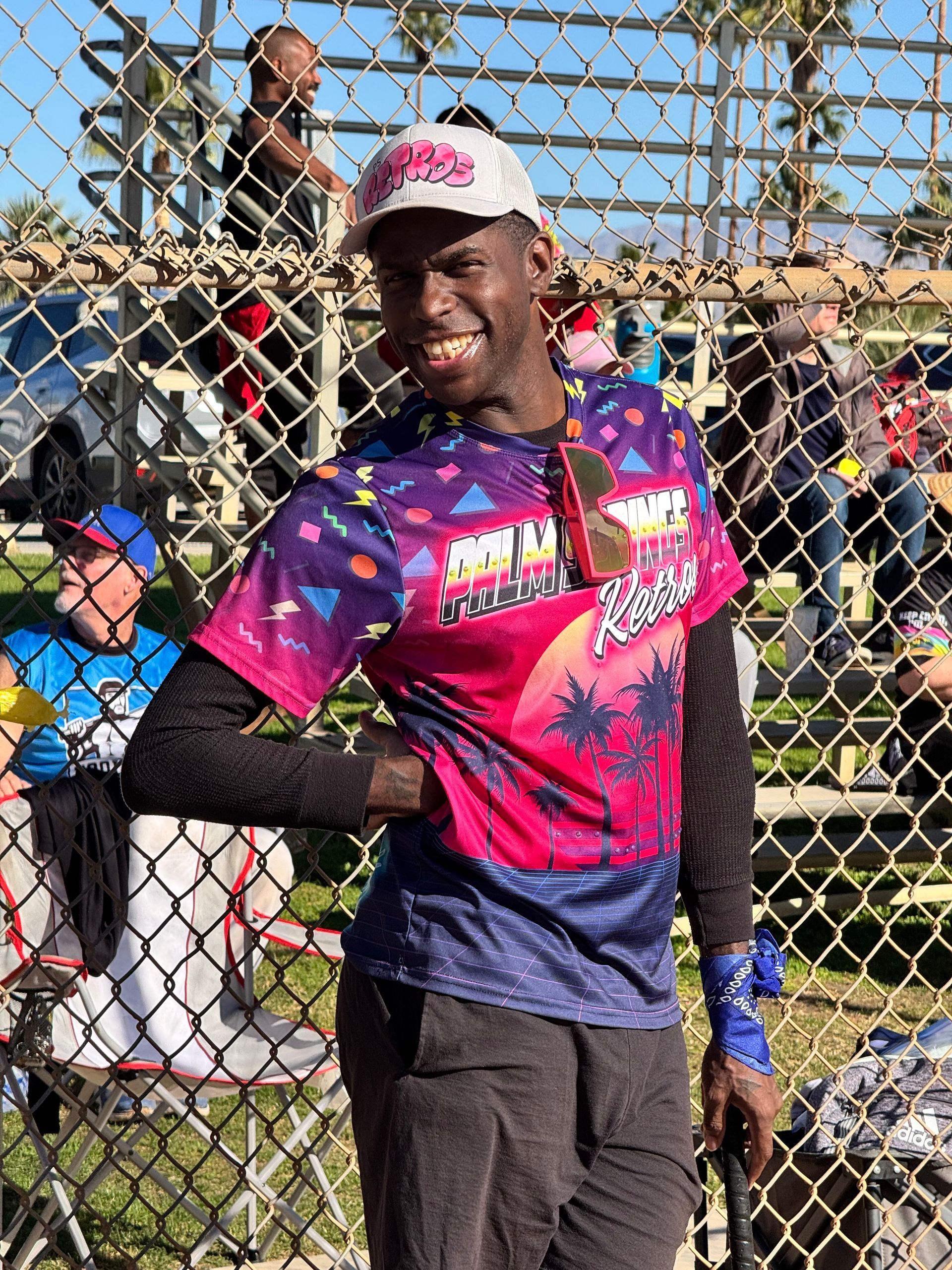 Smiling athlete in pink and purple shirt posing by a chain-link fence on a sunny field