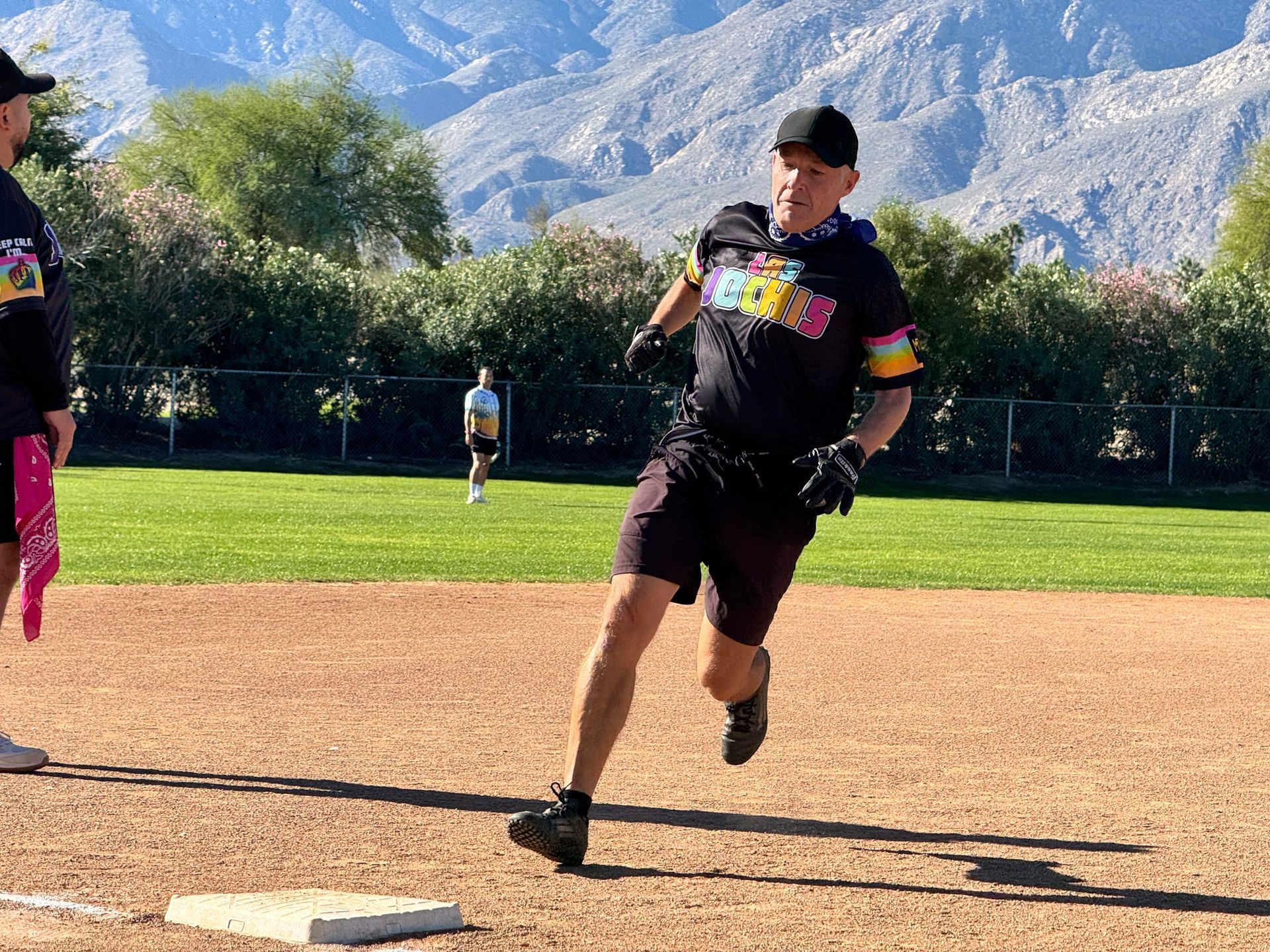 Runner in black shirt and shorts nearing a base on a baseball field with mountains in the background