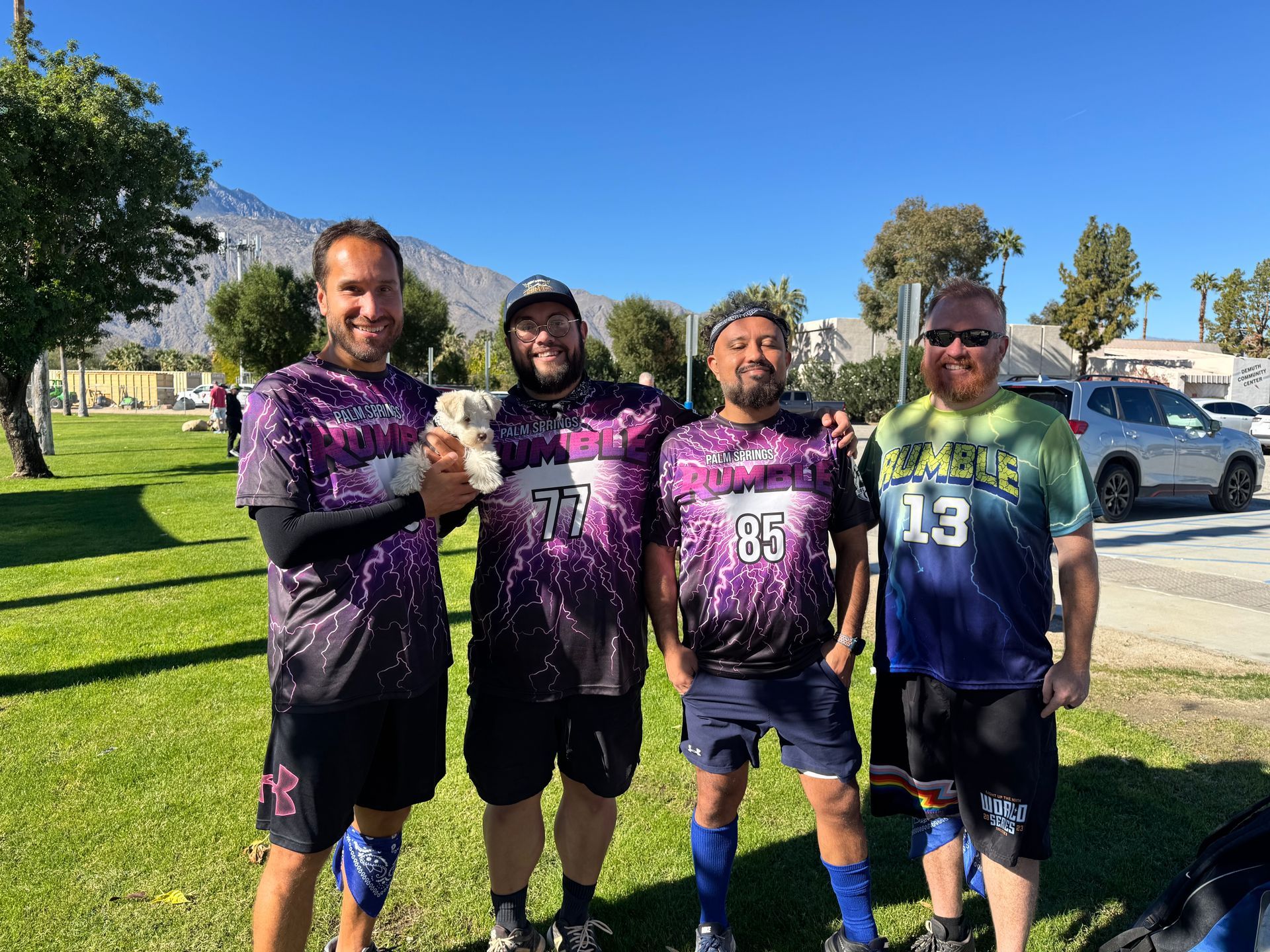 Four athletes in purple jerseys posing on a grassy field with mountains and parked cars behind them