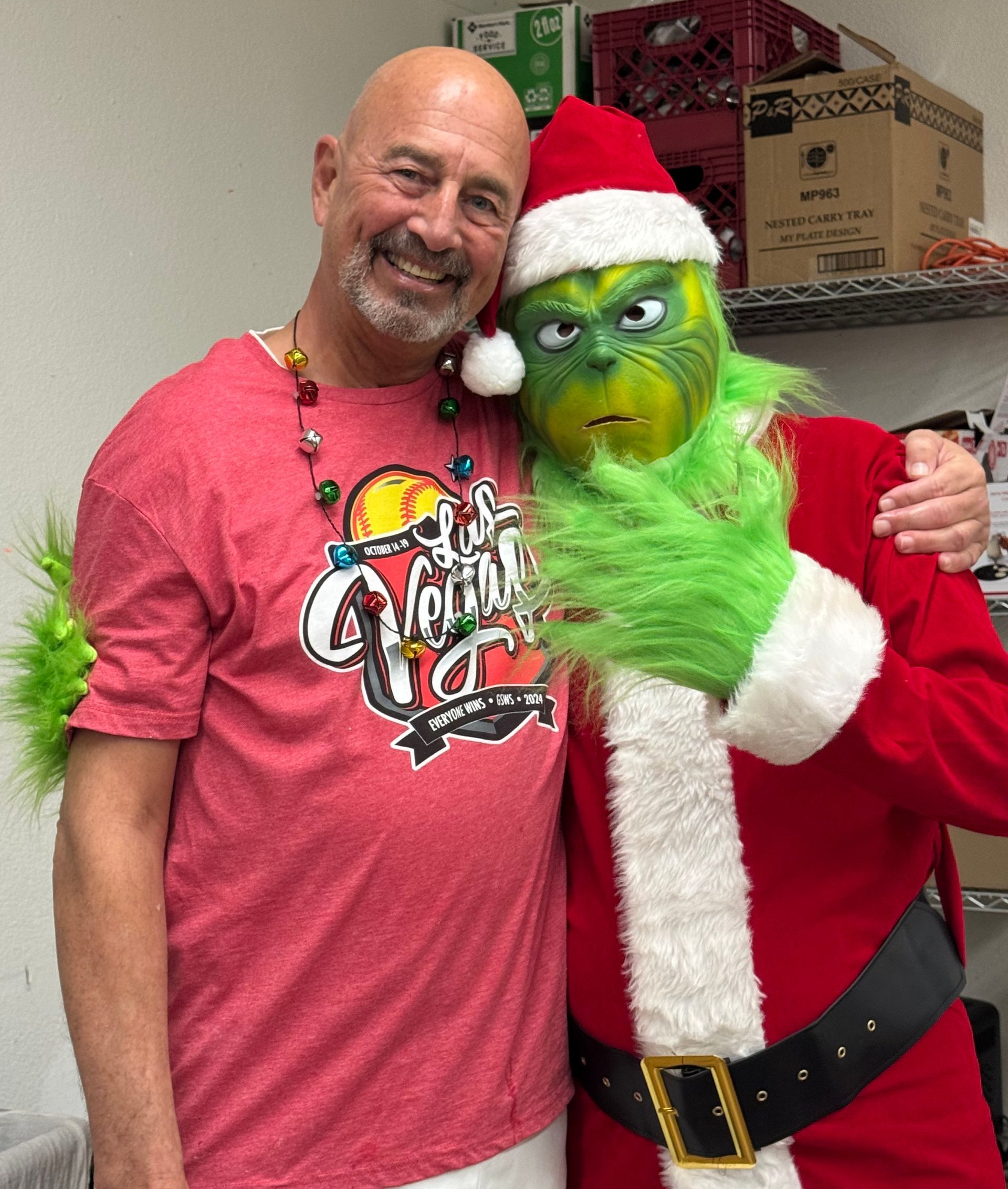 Man in red shirt posing with the Grinch in a Santa outfit in a room with shelves and boxes.
