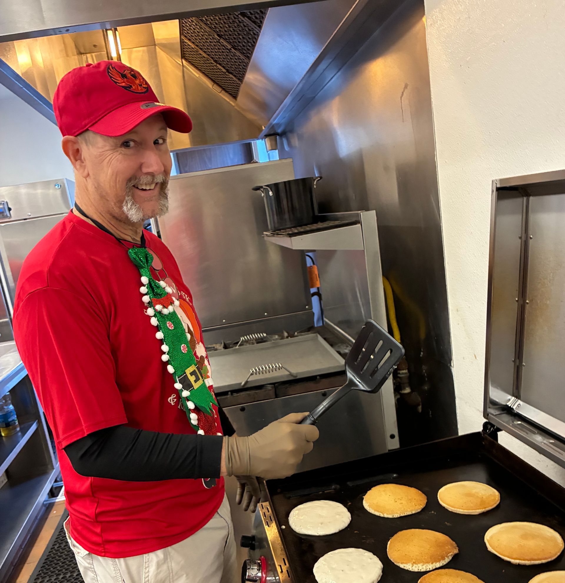 Person in red shirt and cap smiling while flipping pancakes on a griddle in a commercial kitchen