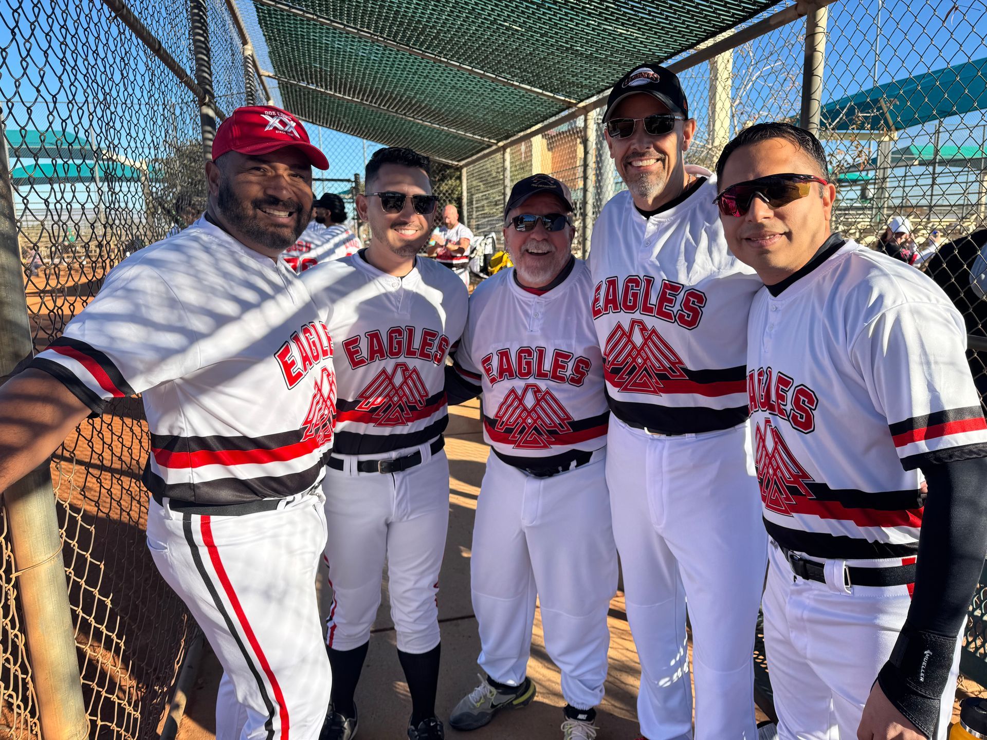 Five baseball players in white uniforms posing together on a field behind a chain-link fence.