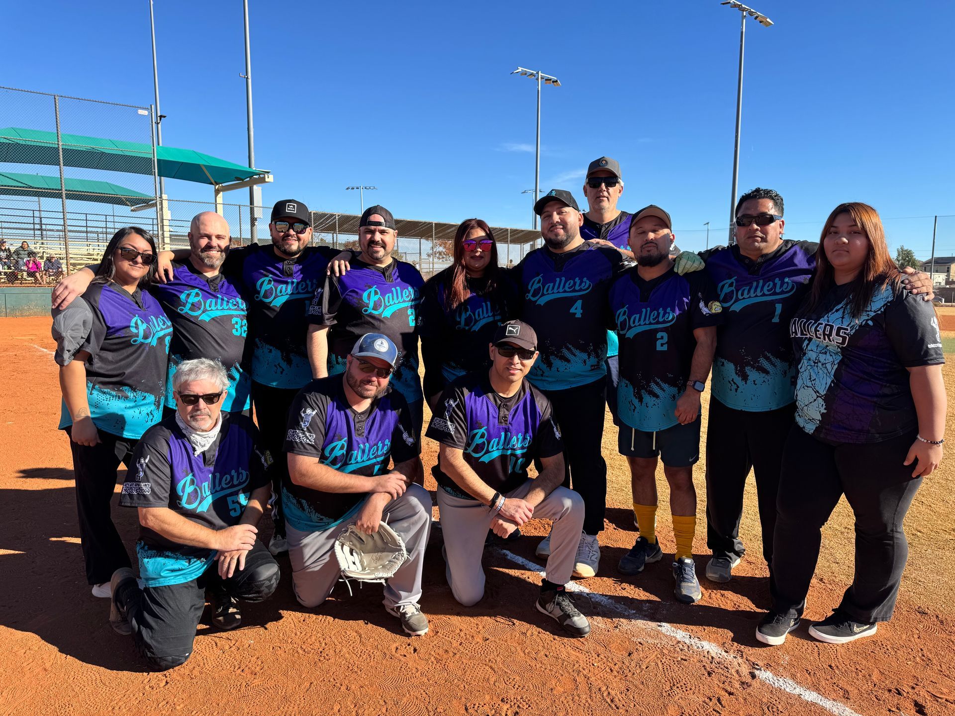 Softball team posing on a dirt field in matching blue and black uniforms under a bright sky