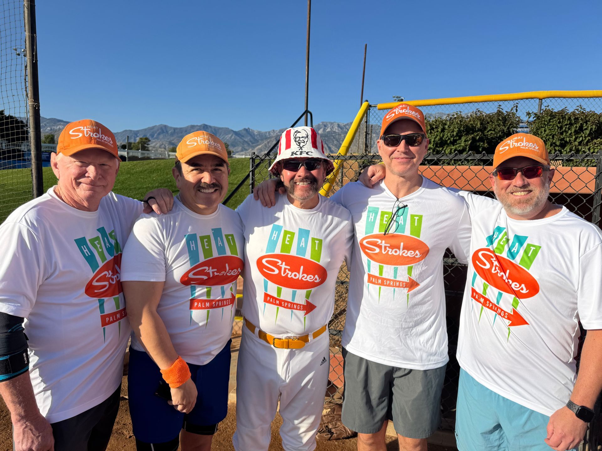 Five people in white shirts and orange caps pose outdoors in front of a scenic mountain view.