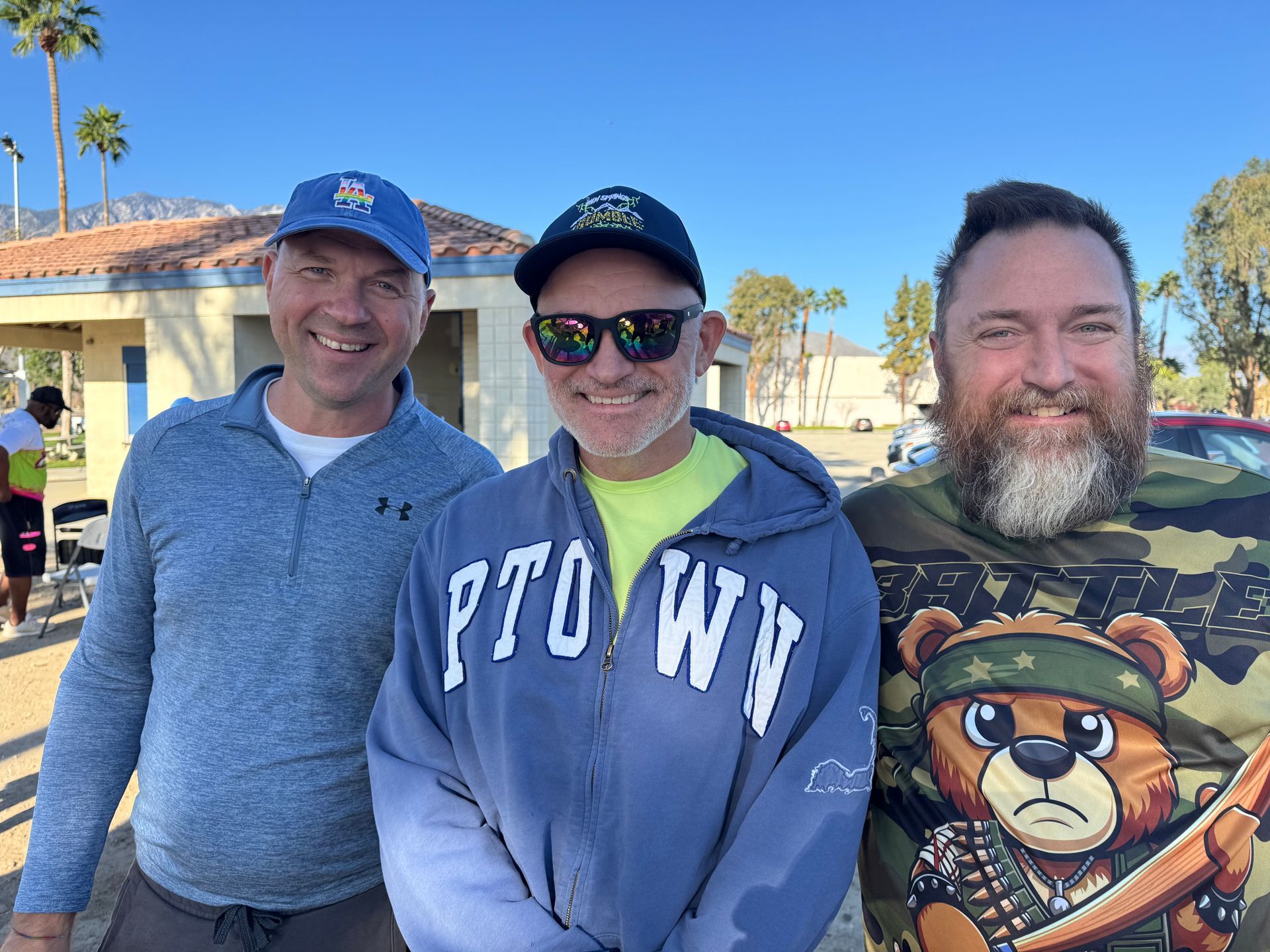 Three smiling people posing outdoors on a sunny day, with houses and palm trees in the background.