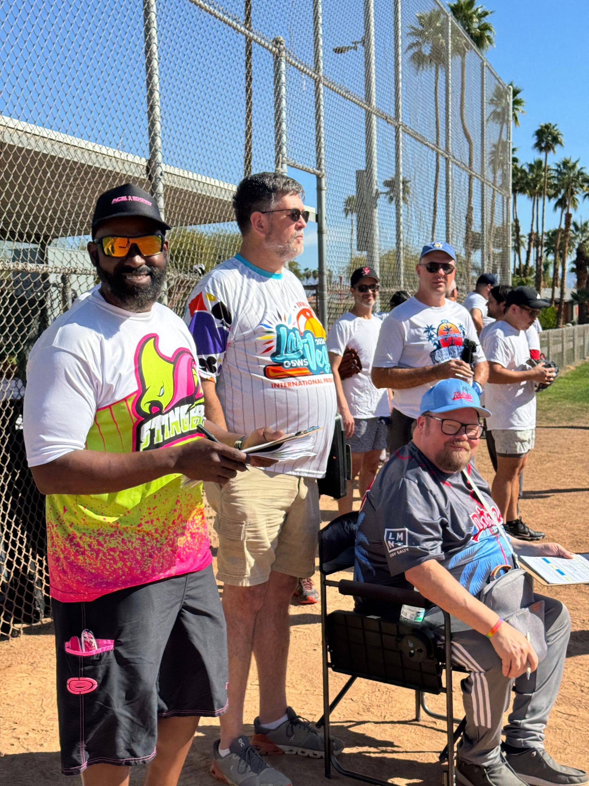 Group of people posing outdoors at a sunny sports court, holding drinks and wearing colorful shirts.