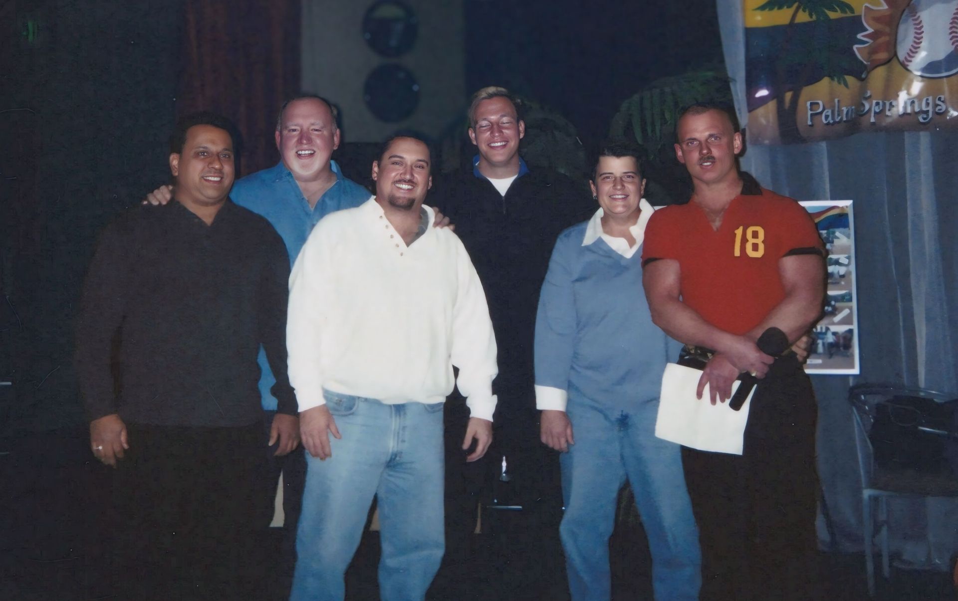 Six people posing indoors, smiling, with one holding a shirt and papers, under dim lighting.