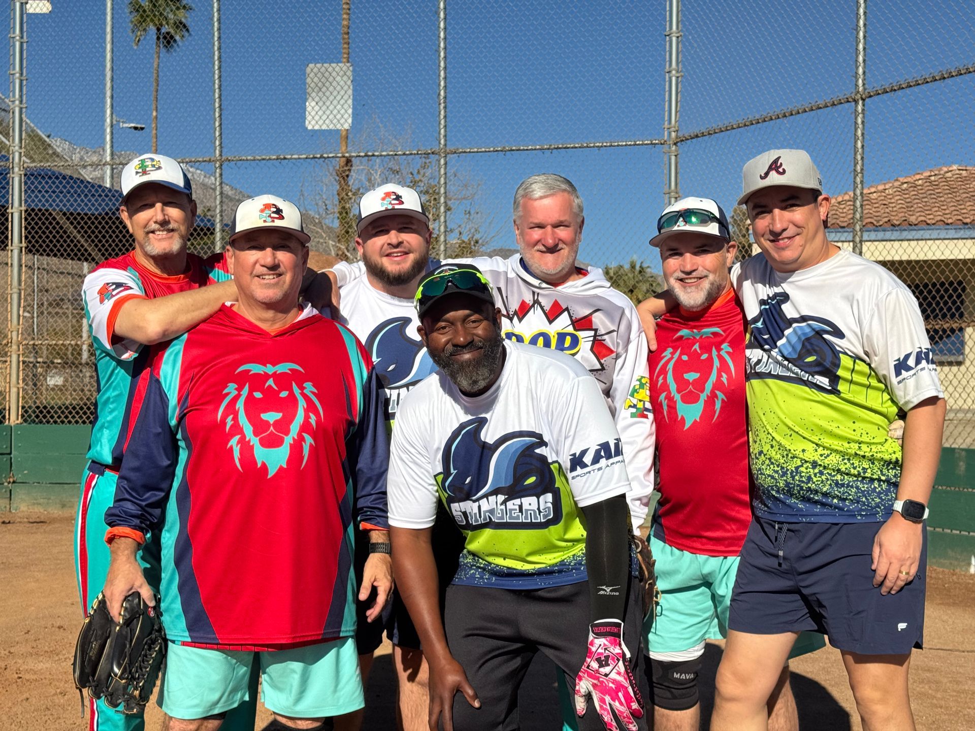 Six men in matching softball uniforms pose together on a sunny field with a chain-link backstop.