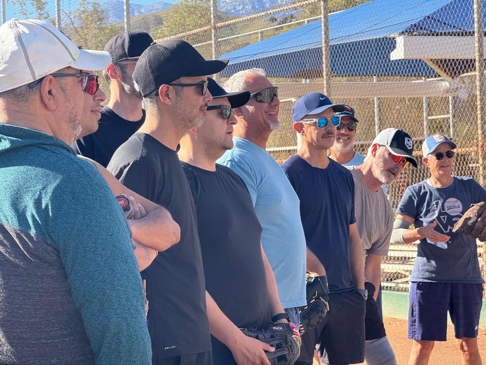 People standing outdoors behind a fence, wearing casual clothes and sunglasses on a sunny day.