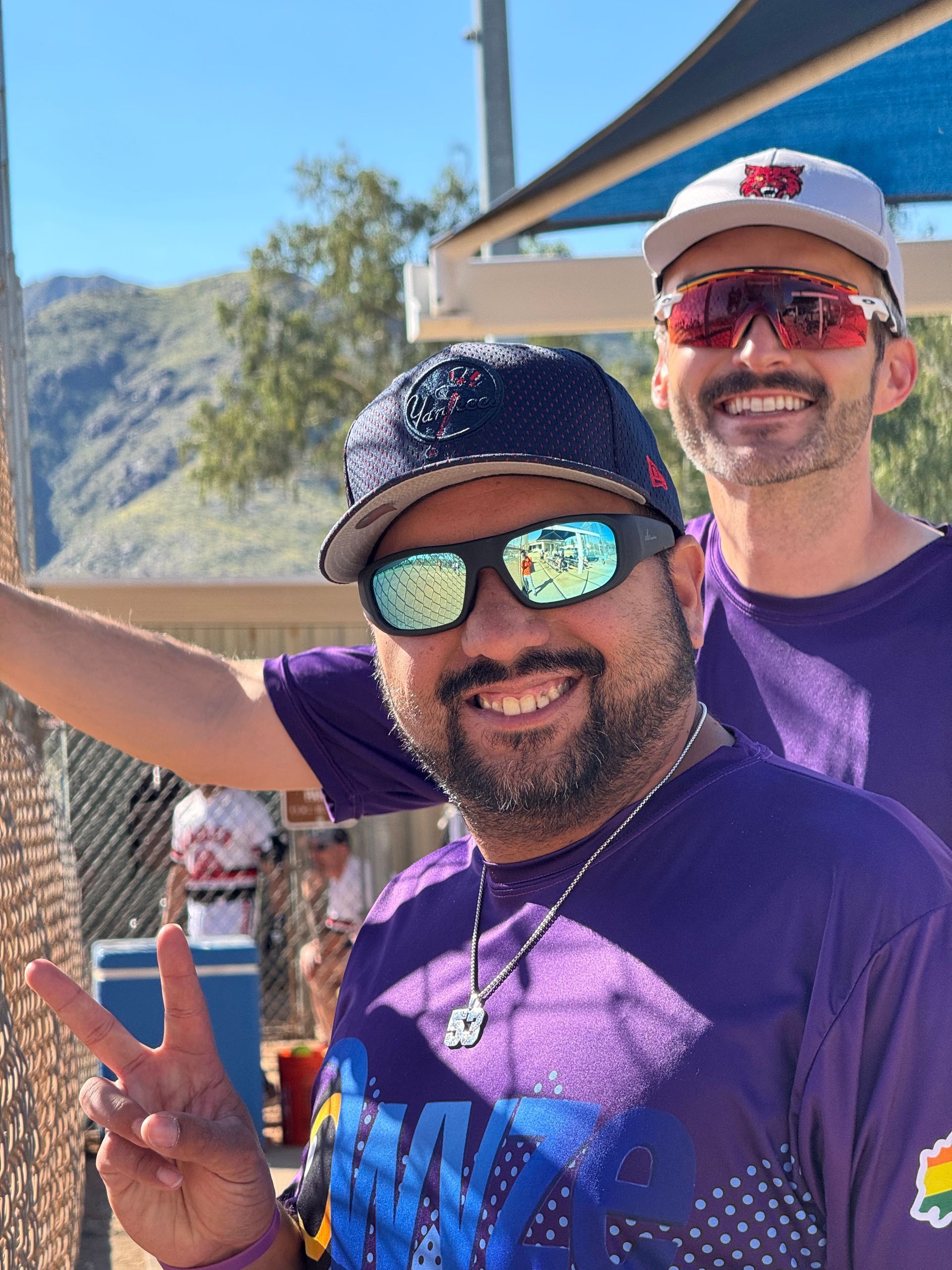 Two people smiling outdoors, one making a peace sign in sunglasses and a purple shirt