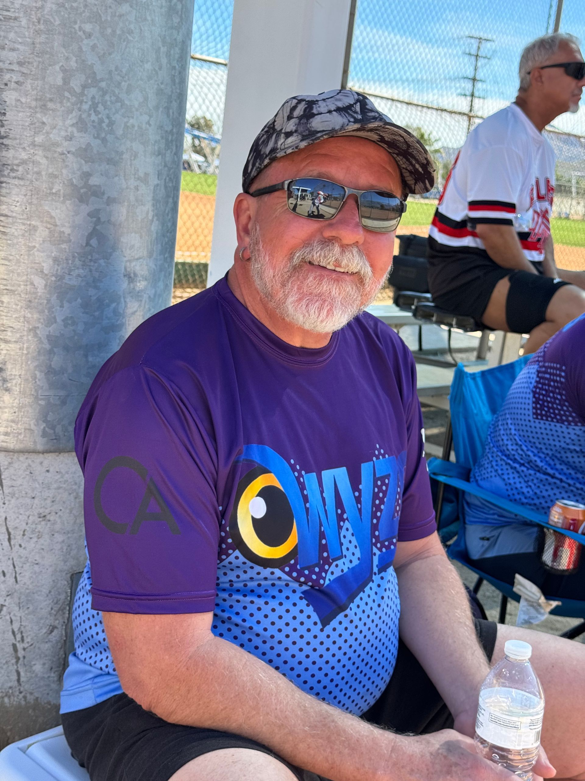 Smiling person in purple shirt and camo cap sitting outdoors at a sporting event