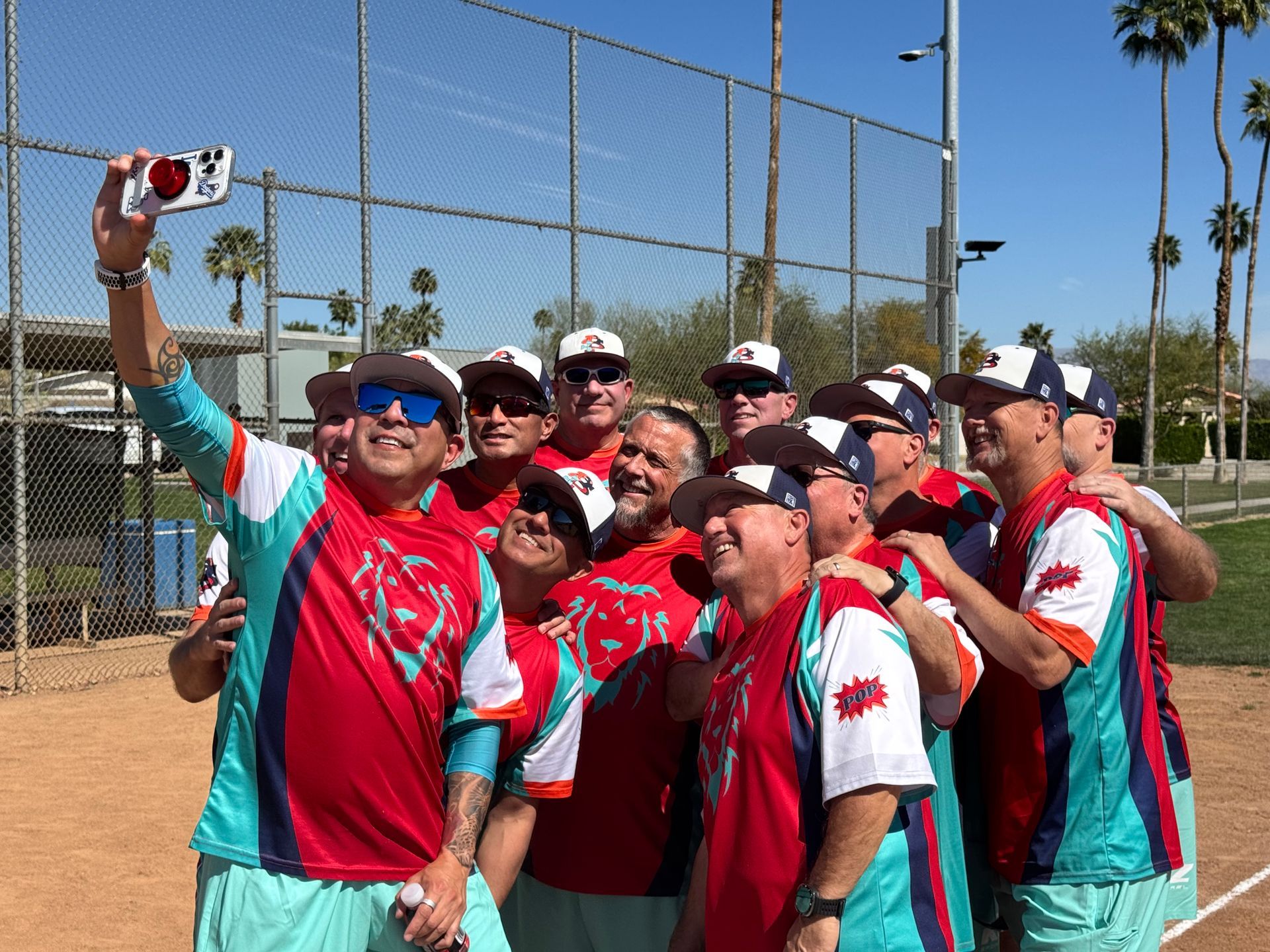 Group of softball players in red and teal jerseys taking a selfie on a sunny field.
