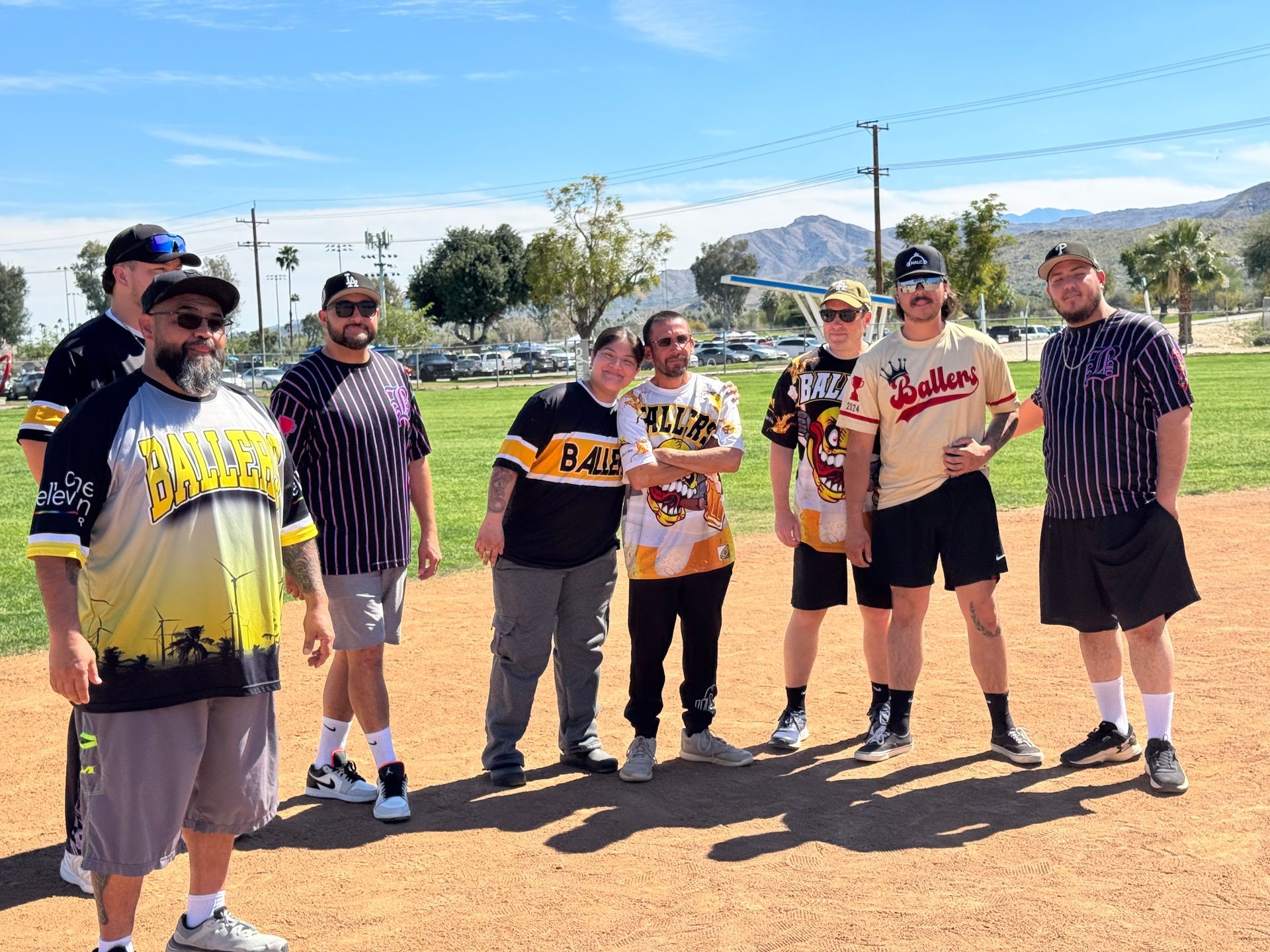 Seven people standing together outdoors in baseball jerseys on a dusty field under a clear sky