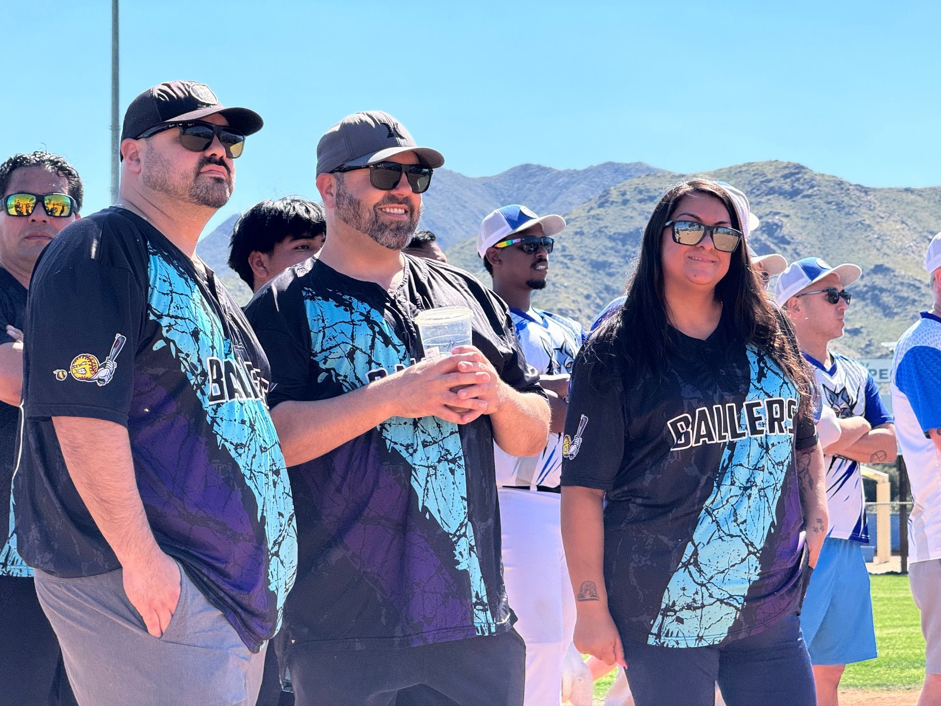 Three people in matching blue-and-black shirts stand outdoors in bright sun, with mountains in the background.