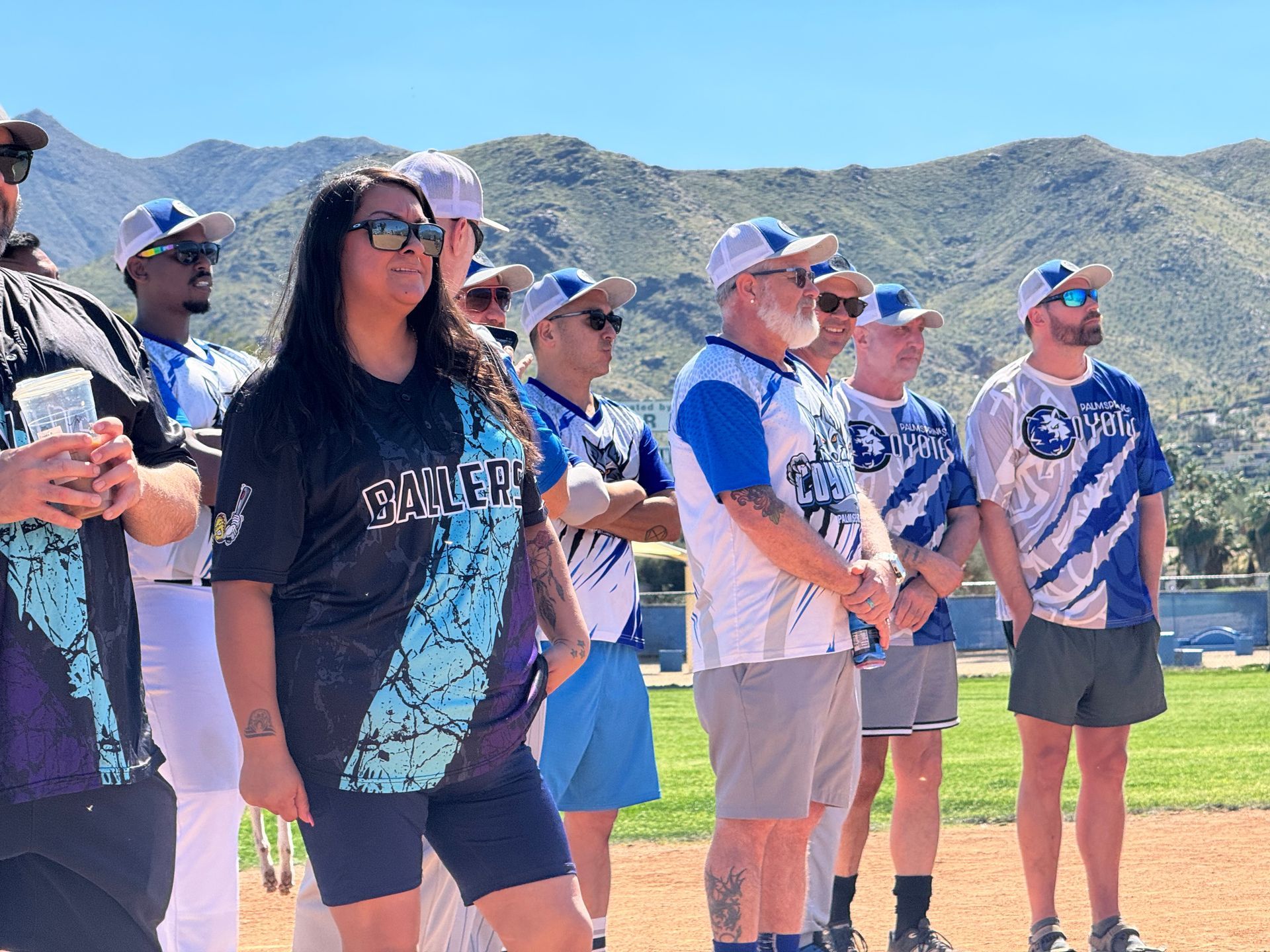 People in matching blue shirts and caps stand on a baseball field with mountains in the background.
