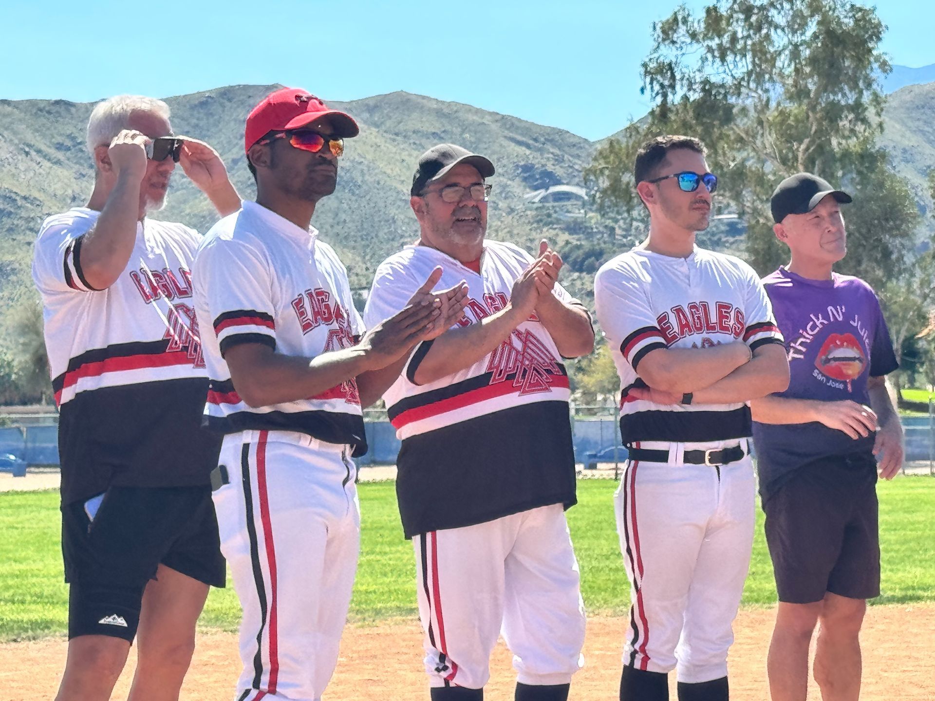 Five men in baseball uniforms stand outdoors, clapping during a mountain-side sports event.
