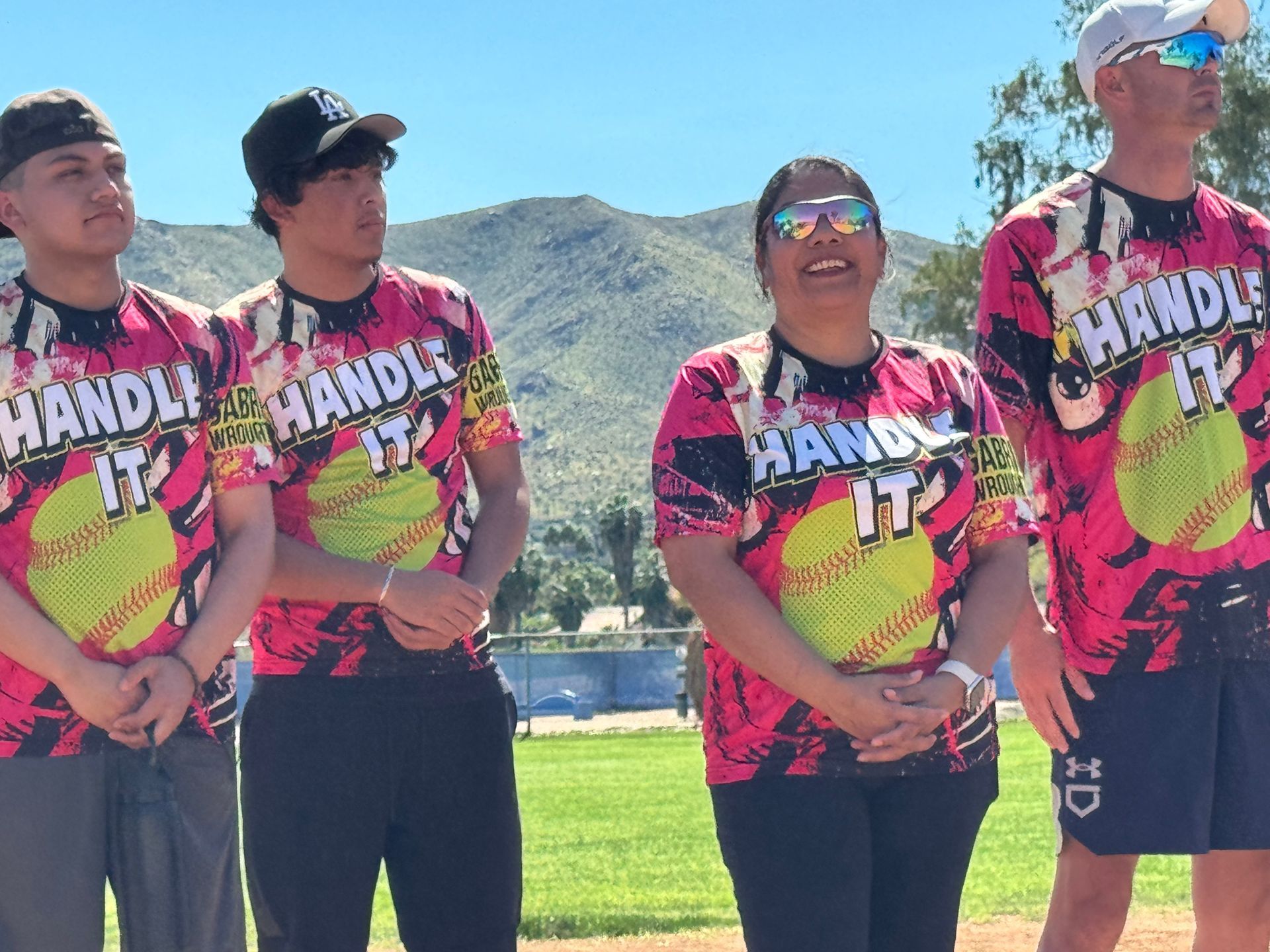 Four people in matching bright shirts and sunglasses standing outdoors in a sunny field with mountains behind them