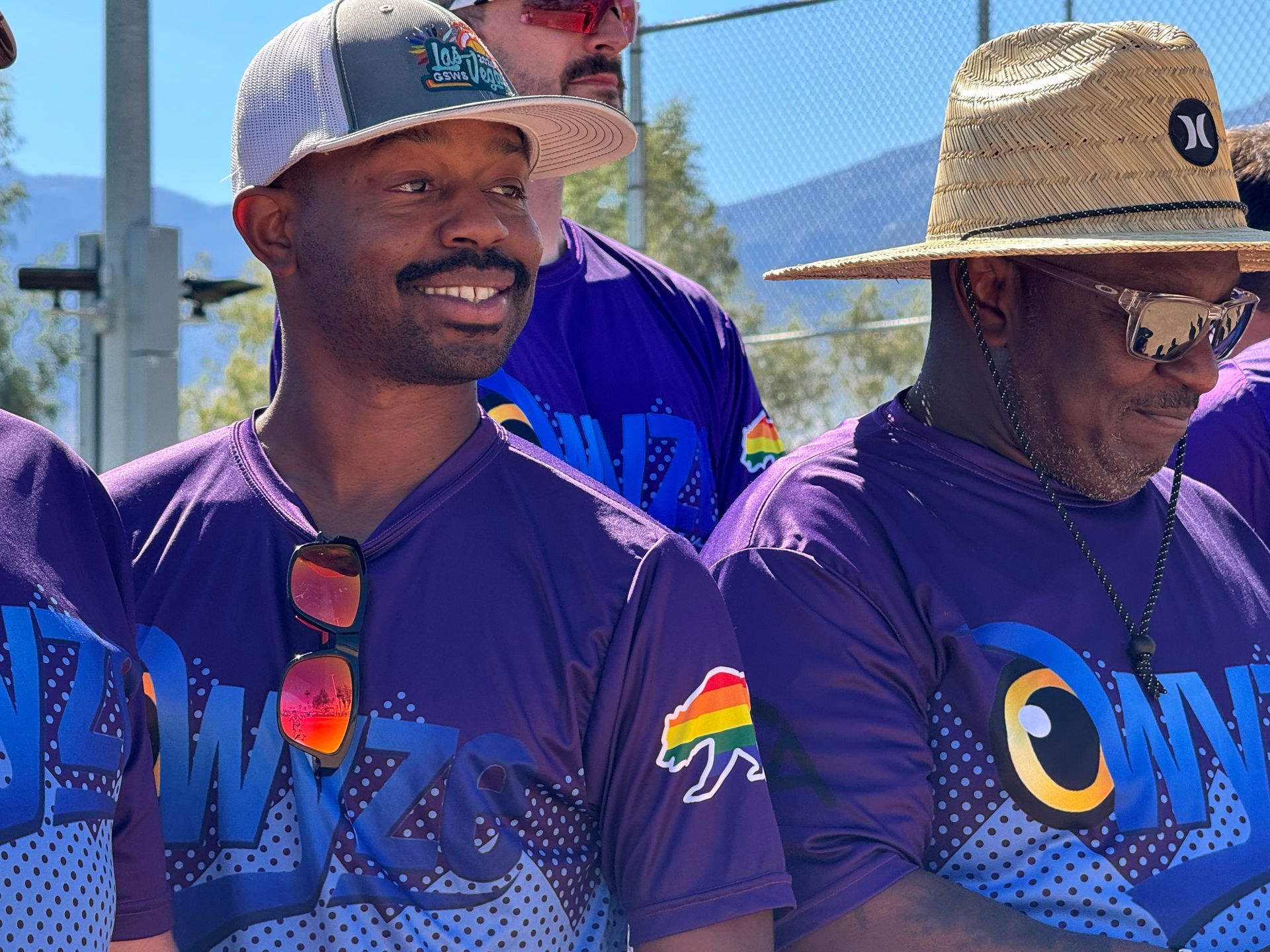 Two men in purple jerseys and hats smiling outdoors, with mountains and a bright sky behind them