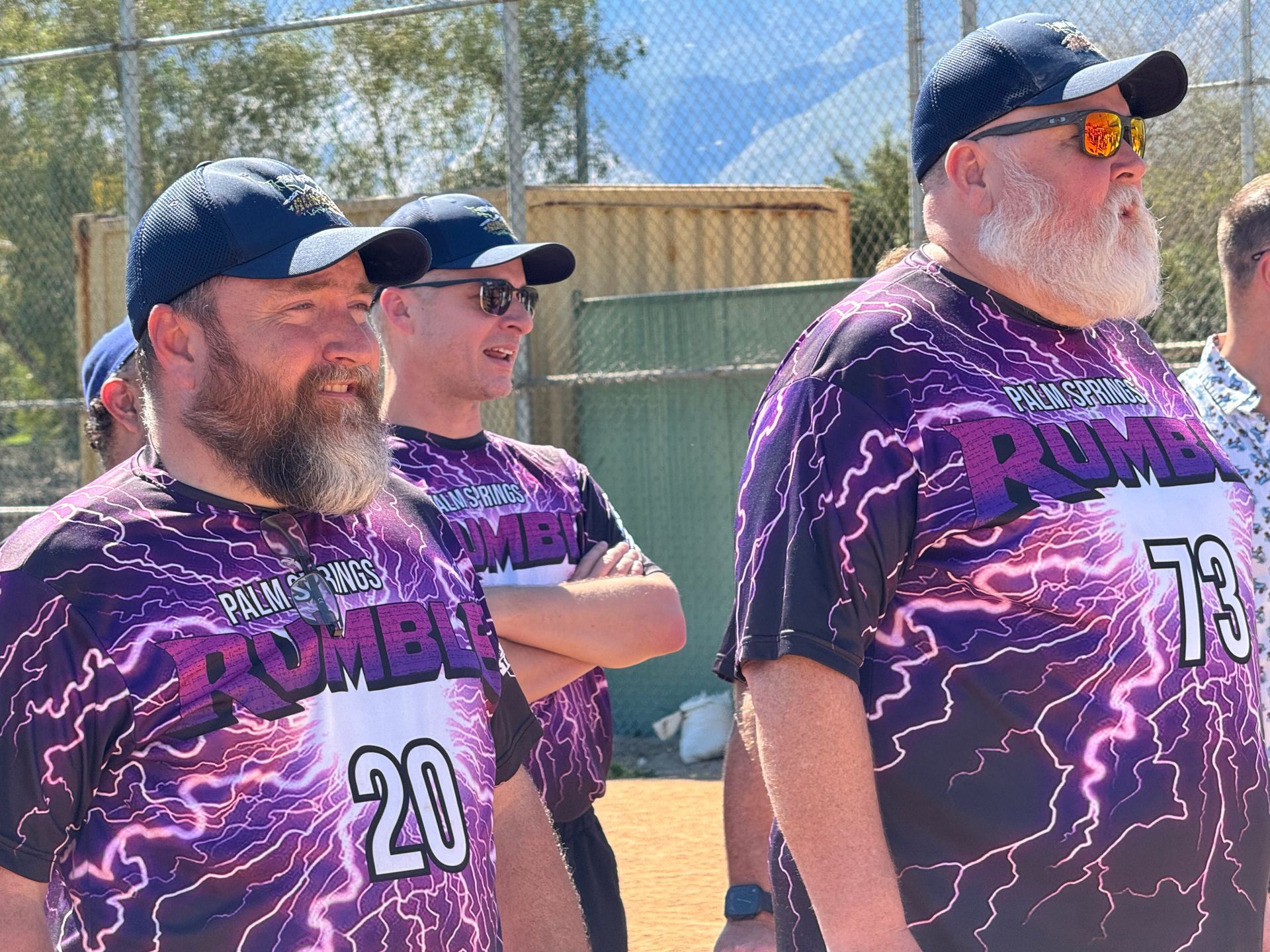 Three people in matching purple jerseys and caps stand outdoors in sunlight.