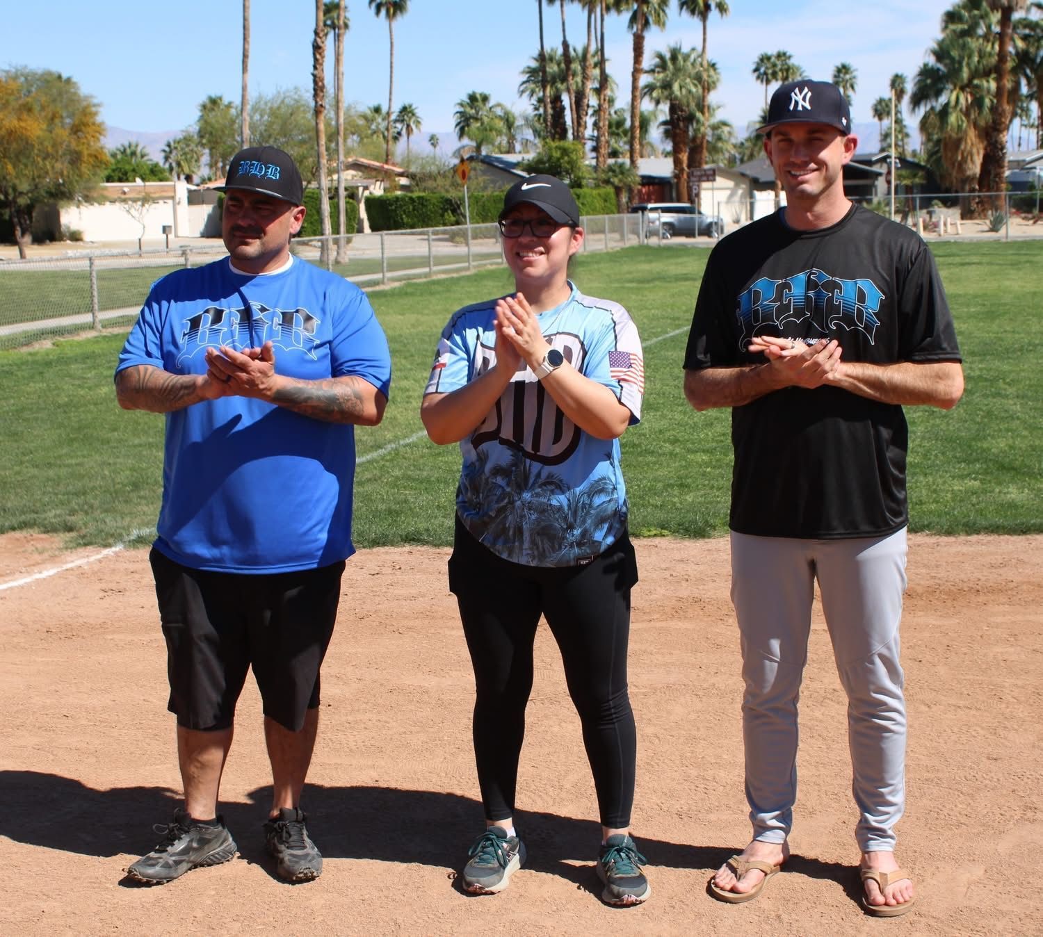 Three people standing on a softball field holding trophy cups in bright daylight