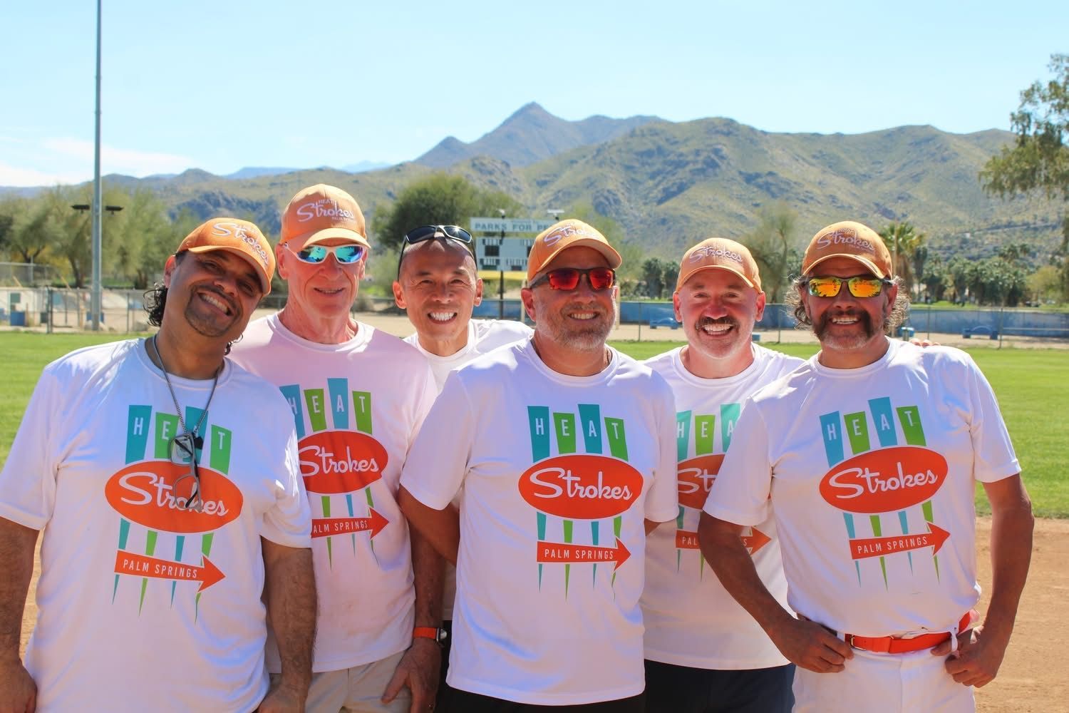 Six smiling runners in white shirts and orange caps standing outdoors with mountains behind them