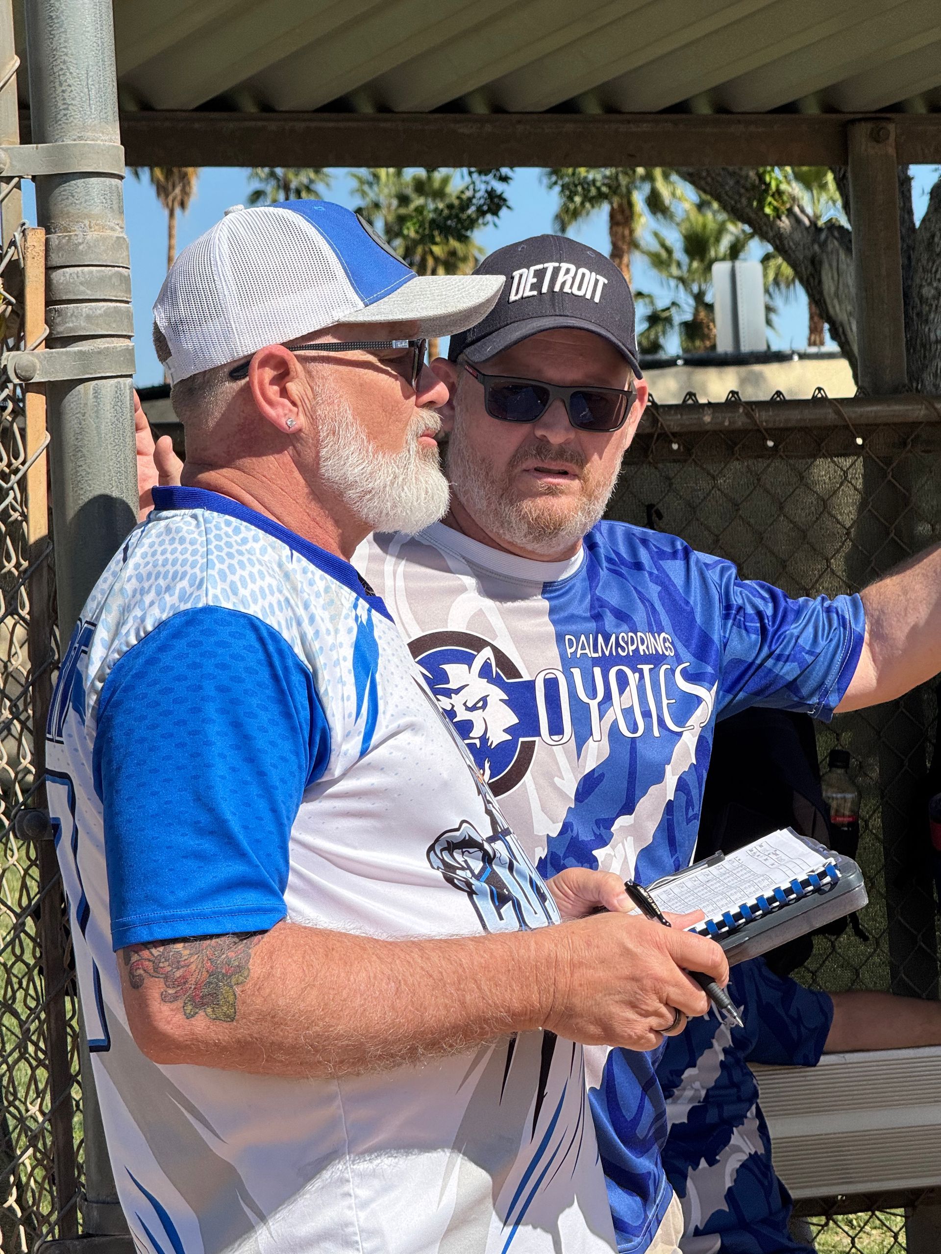 Two men in blue-and-white jerseys chatting under a shaded pavilion, one holding a notebook