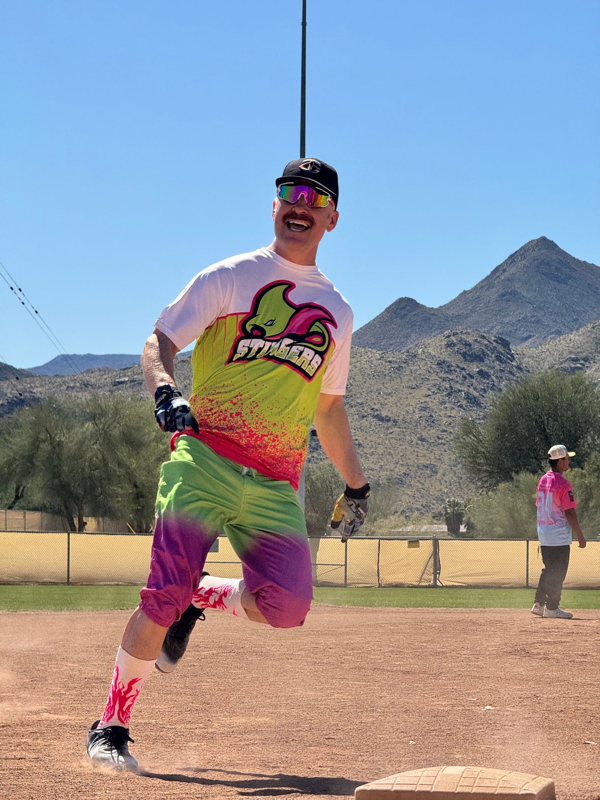 Runner in a colorful outfit mid-stride on a dusty trail with mountains and clear blue sky behind.