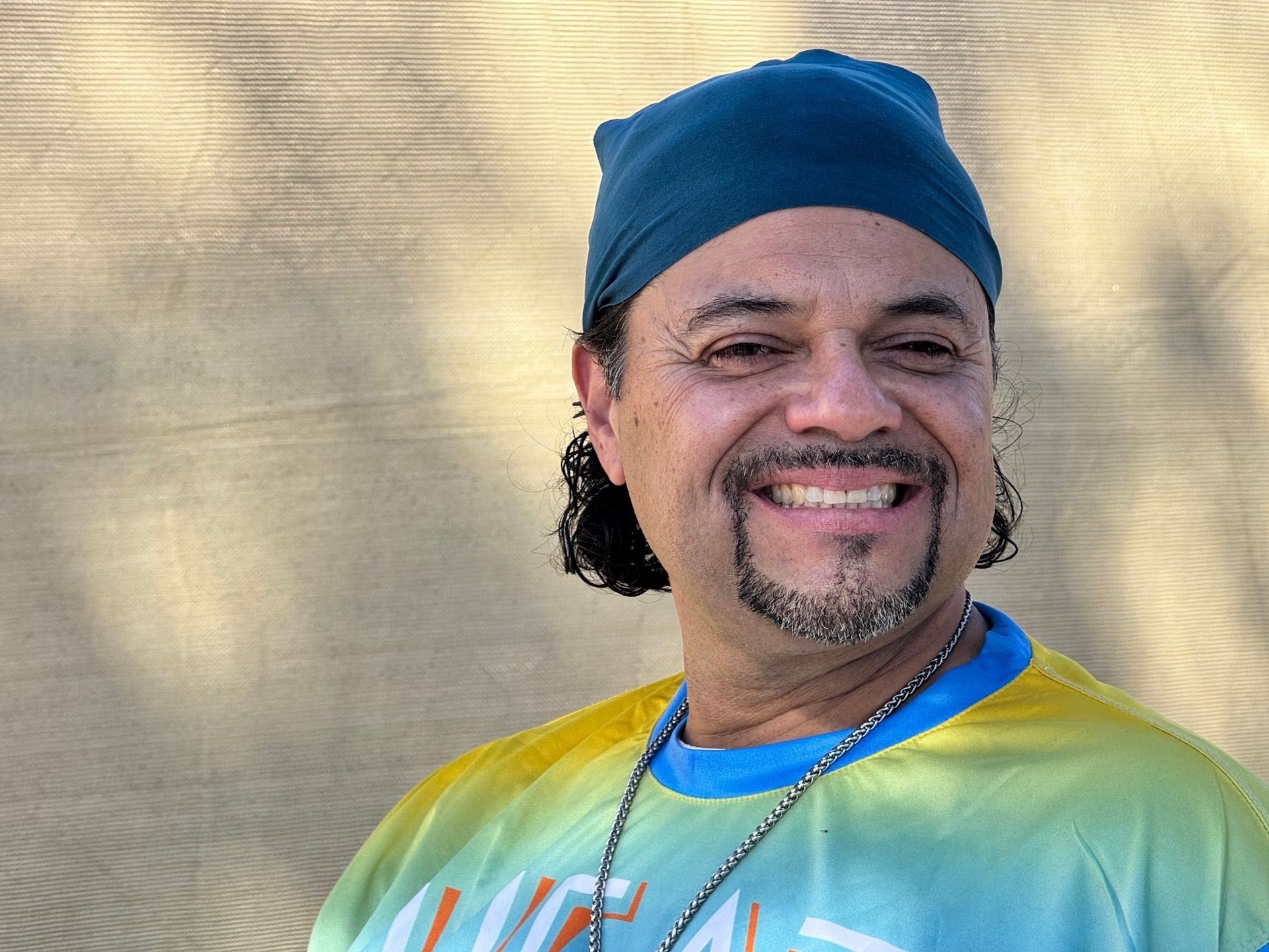 Smiling person in a blue headscarf and rainbow shirt against a beige wall.