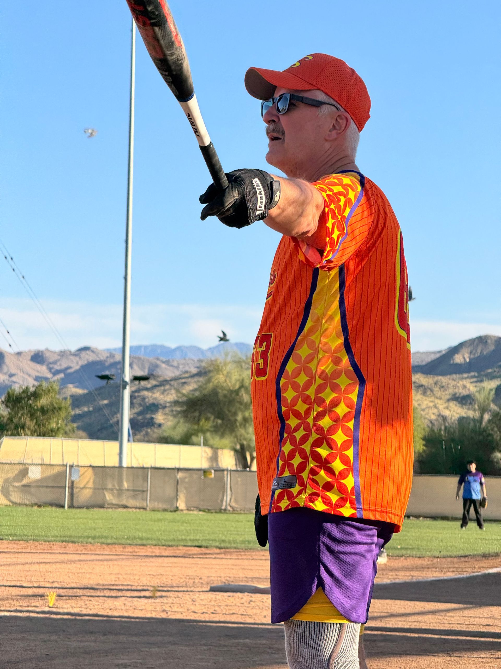 Baseball player in orange jersey and red cap swinging a bat on a field with mountains in the background