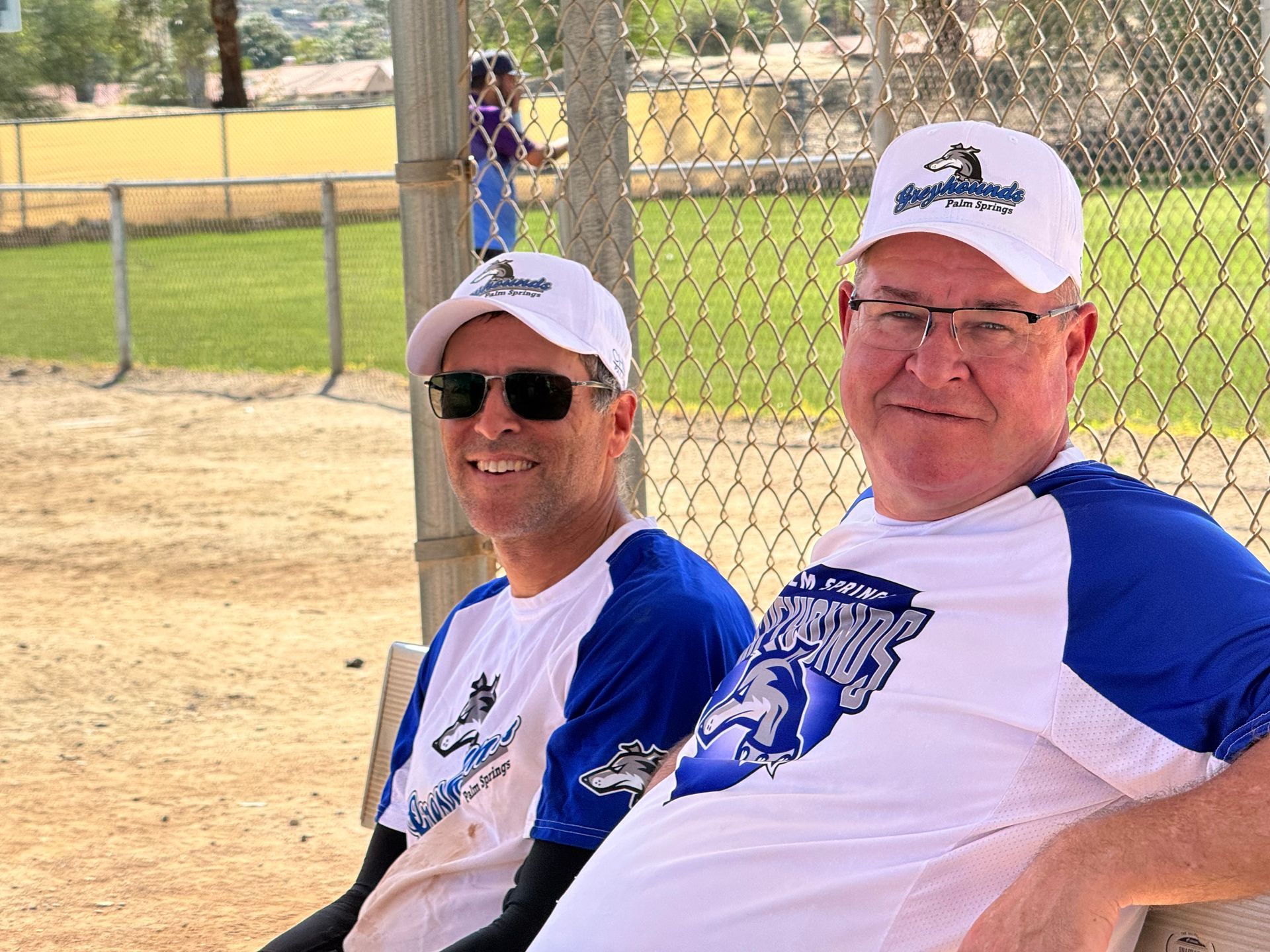 Two people in blue-and-white shirts and white caps sit by a chain-link fence at a baseball field.