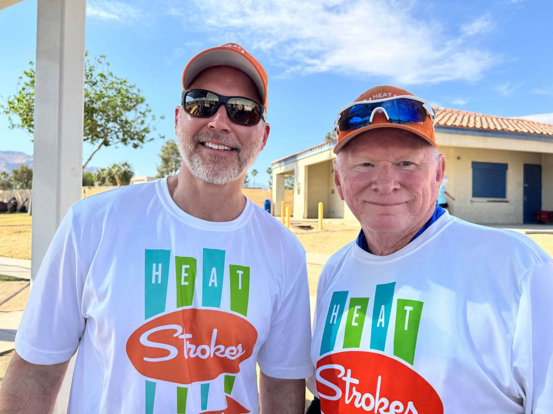 Two smiling people in white T-shirts and orange caps standing outdoors in front of a house.